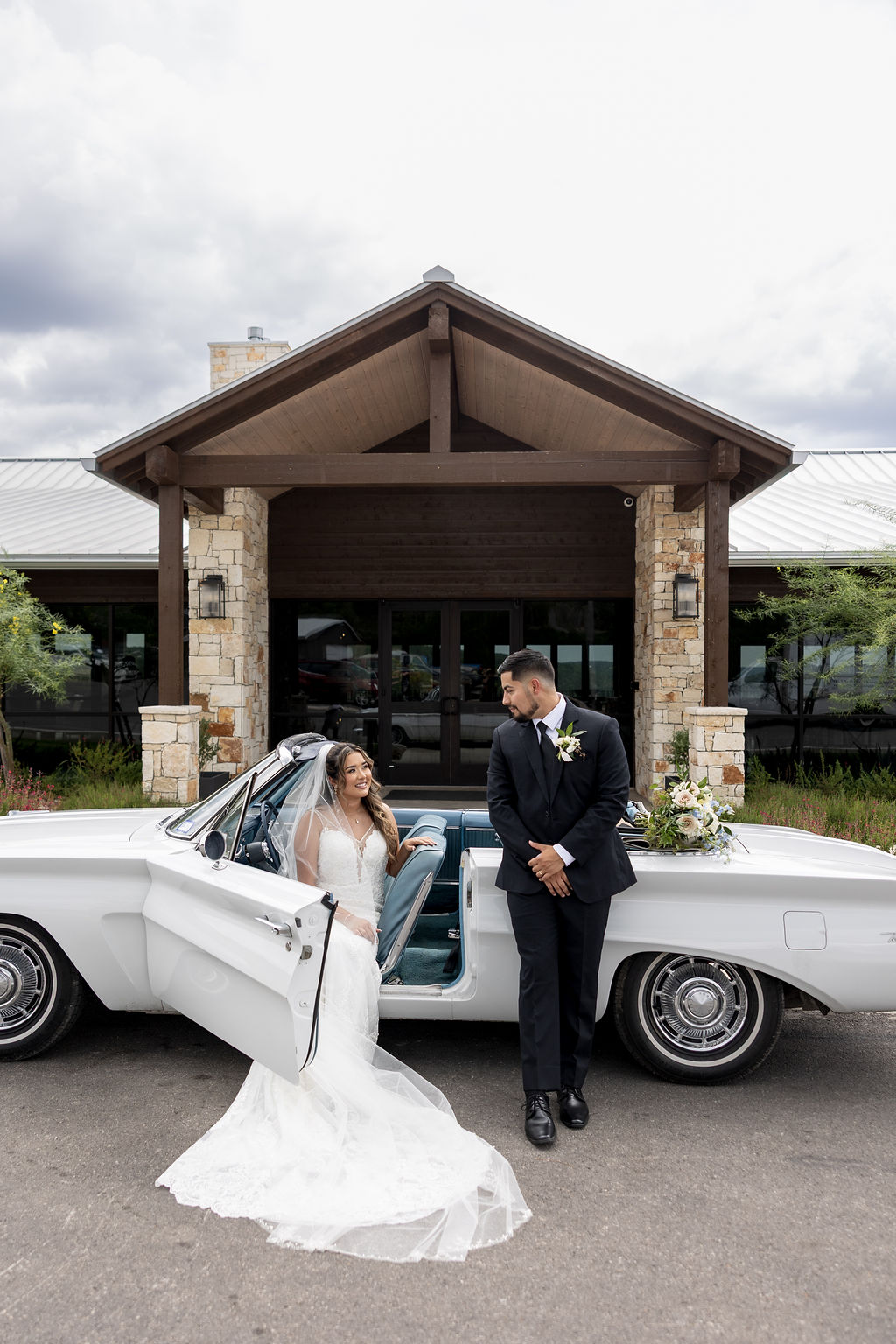 Bride and groom stand outside a building with a vintage white car, bride in a wedding dress, groom in a suit, cloudy sky.