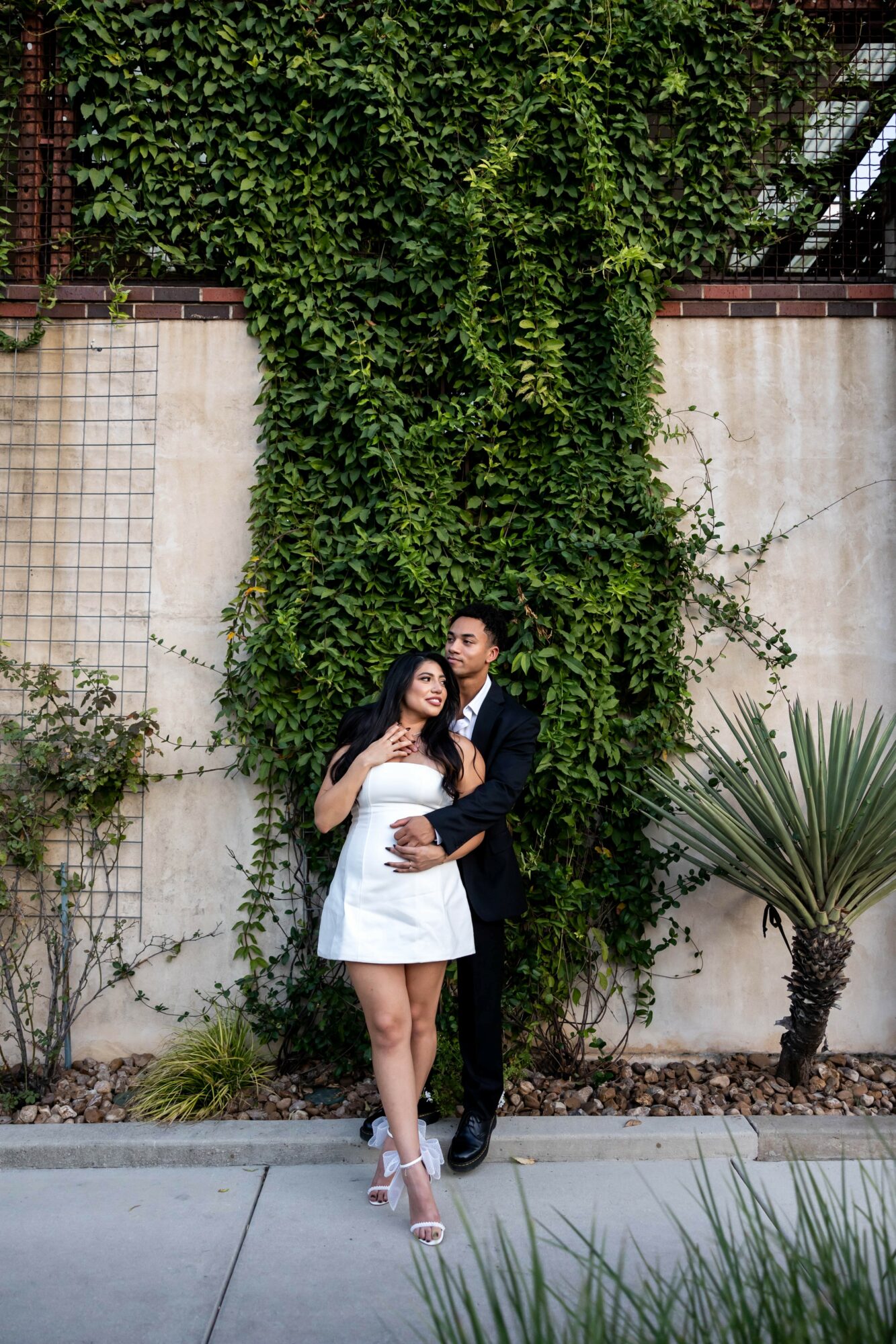 A couple stands in front of a wall covered with green ivy, with plants and a large spiky plant nearby.