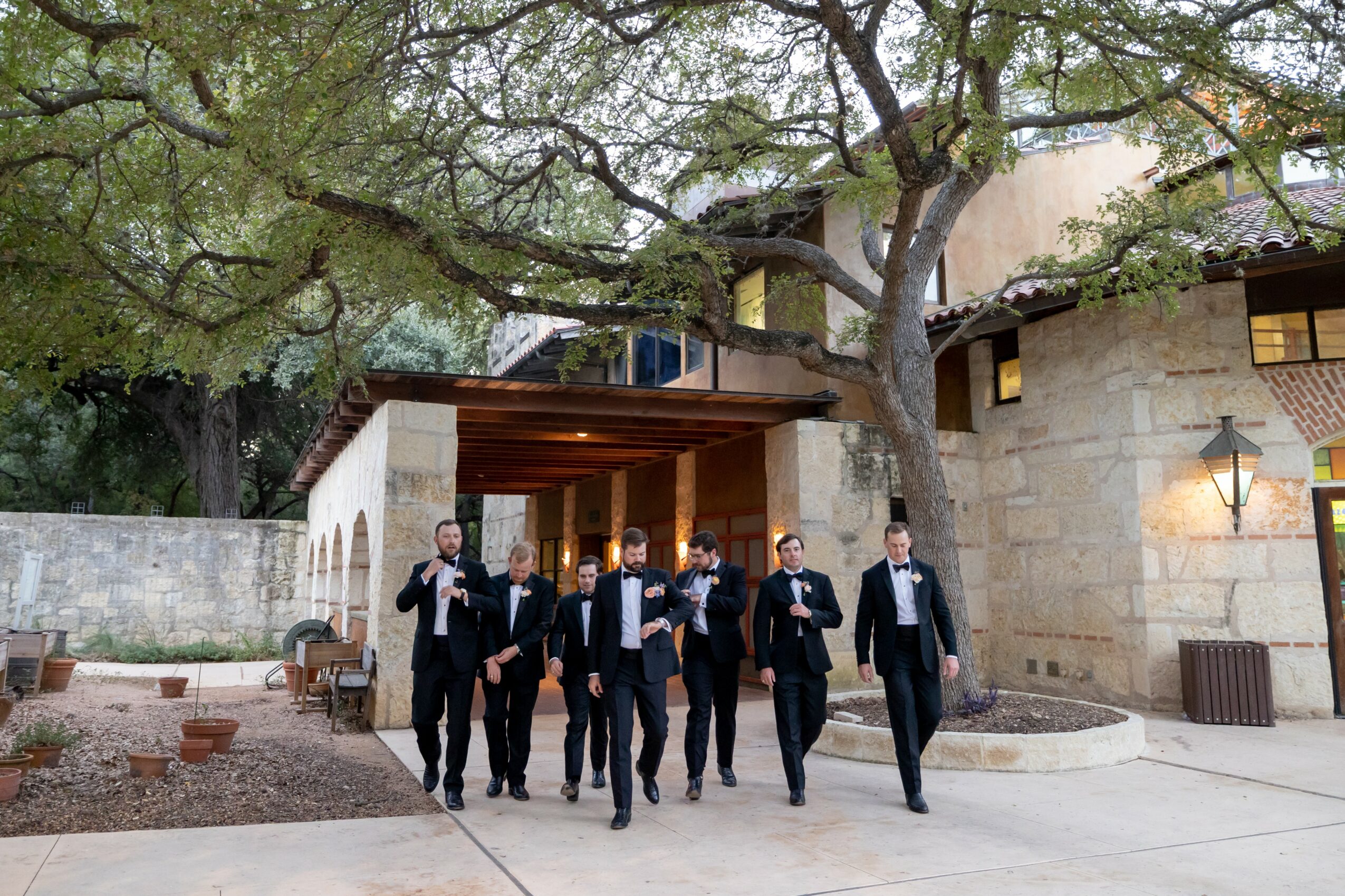 Group of people in formal suits walking outdoors near a stone building and large tree.