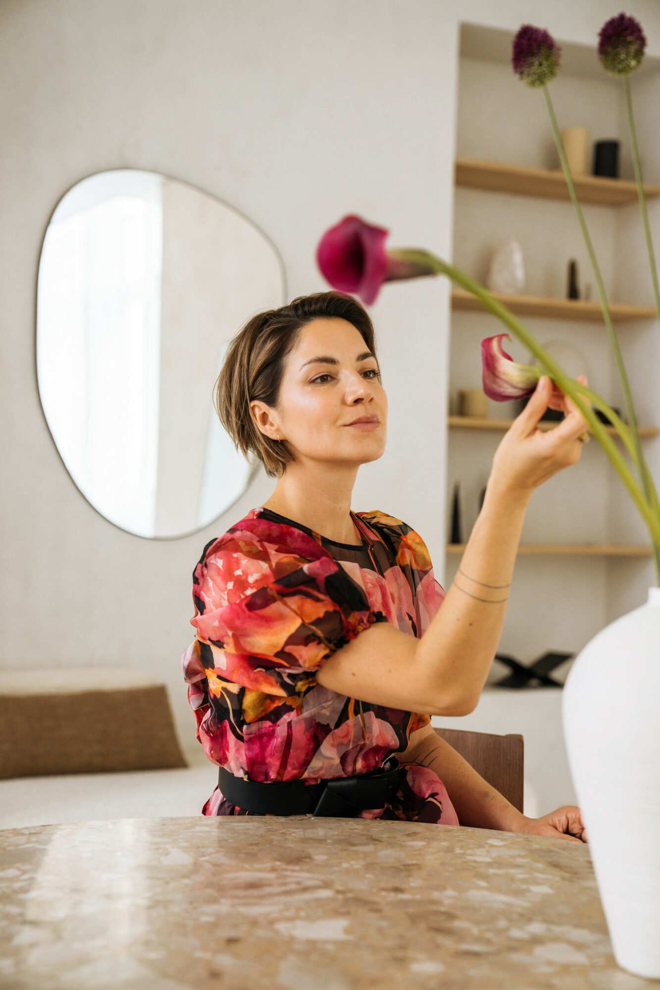 Woman with short hair in a colorful floral top holds a pink flower in a bright room with a mirror and shelves