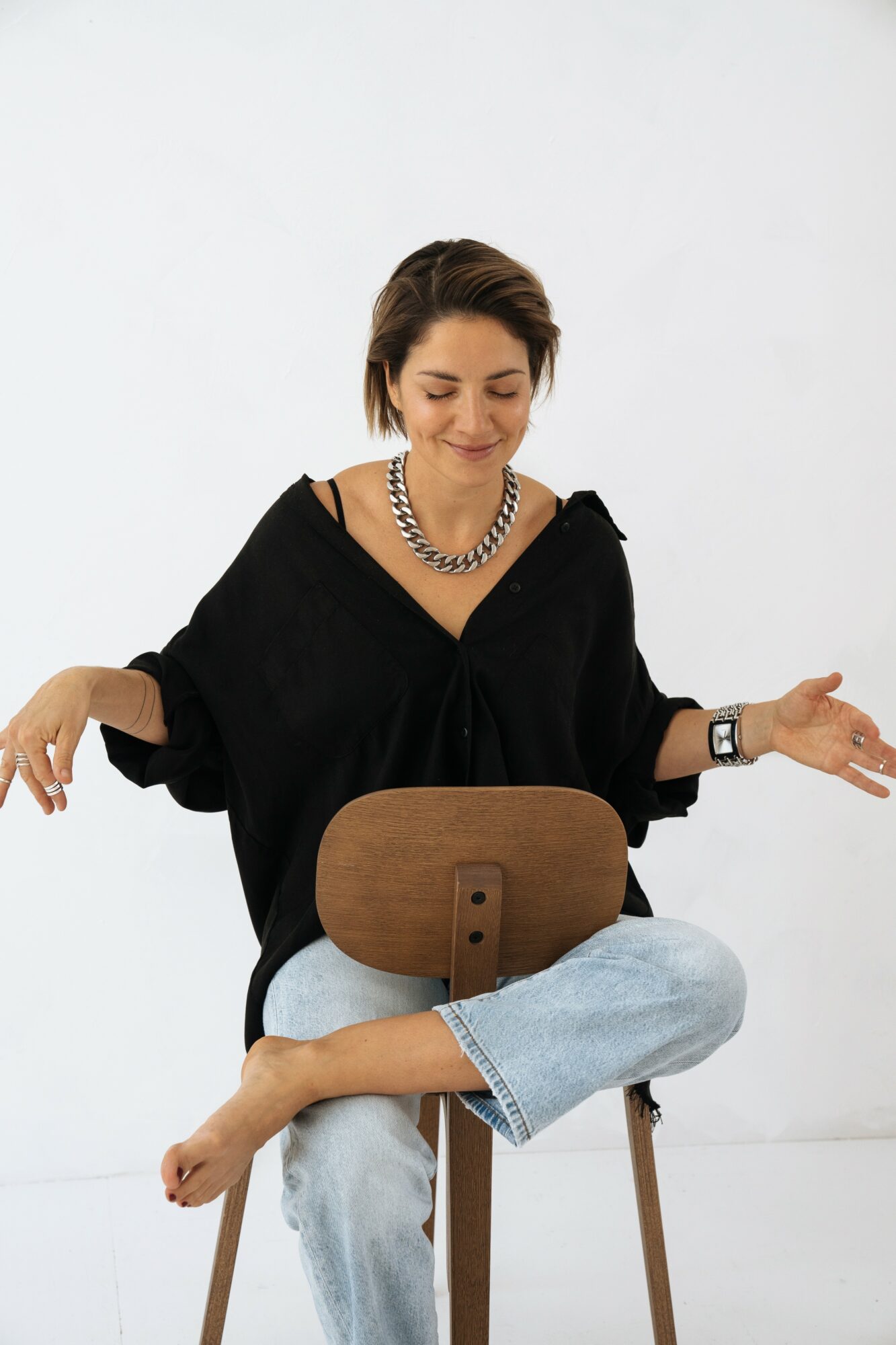 Woman sitting on a chair with crossed legs, smiling, wearing a black top, necklace, and watch, against a white background.