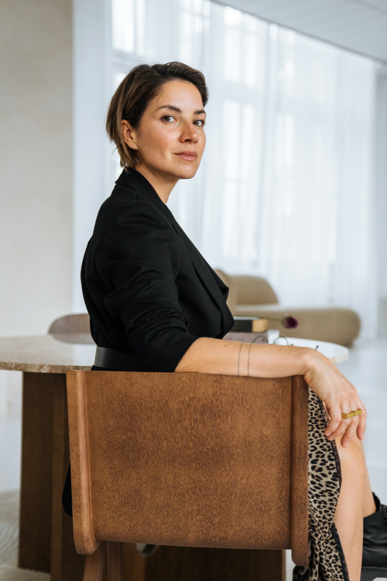 Woman with short hair in a black blazer sitting on a chair in a bright room with large windows.