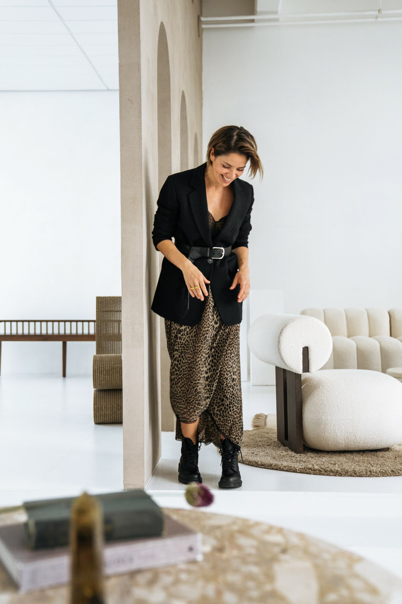 Woman standing indoors near a wall, wearing a black blazer, patterned pants, and black boots, smiling and looking down.