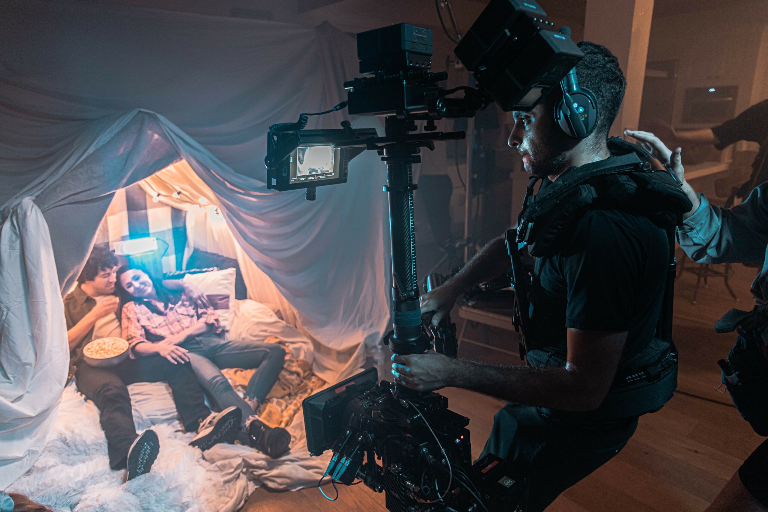 Filmmaker operating camera equipment in a dimly lit room with a person lying on a bed, surrounded by draped fabric.