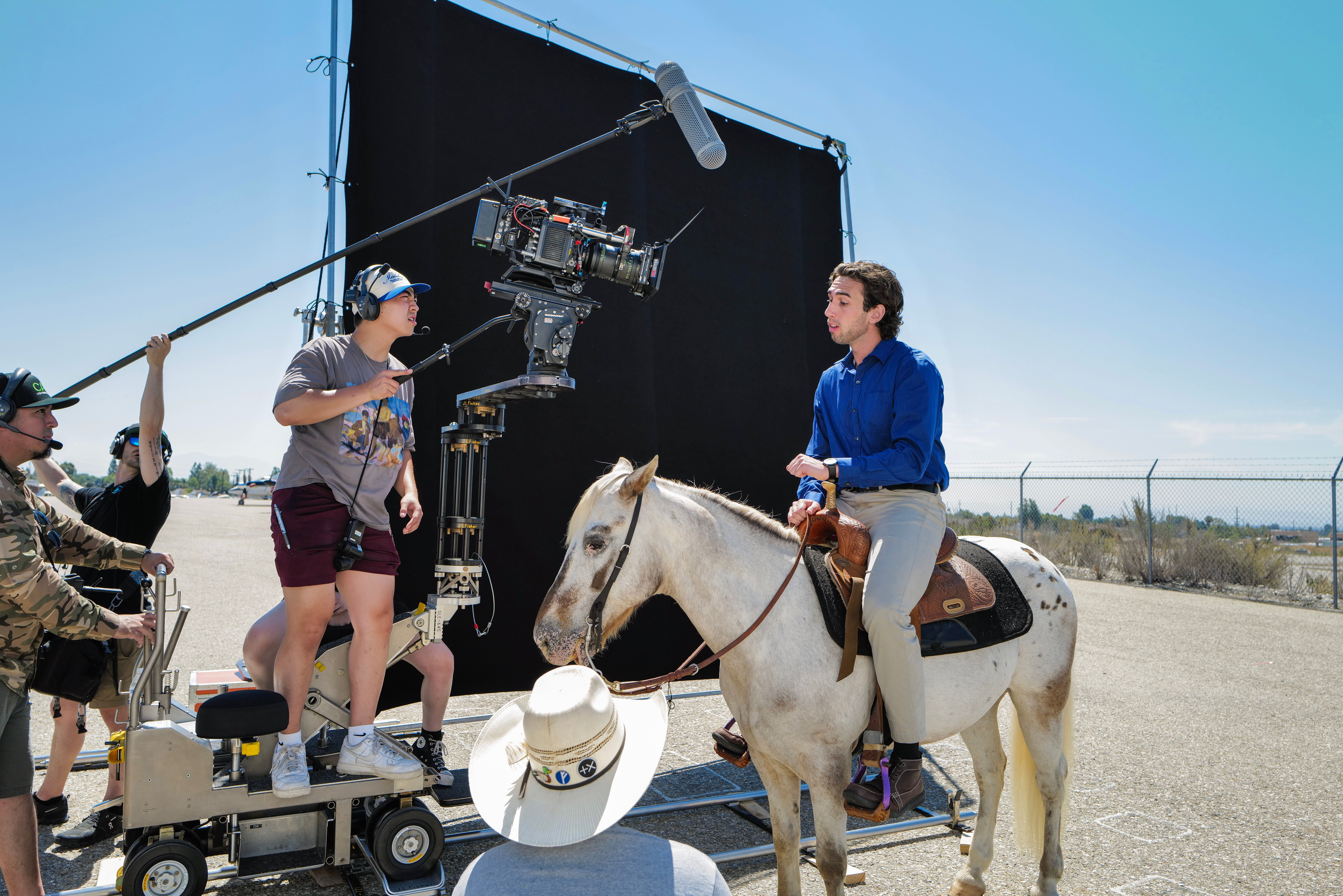 Person riding a white horse on a film set with crew and camera equipment, outdoors under a clear sky.