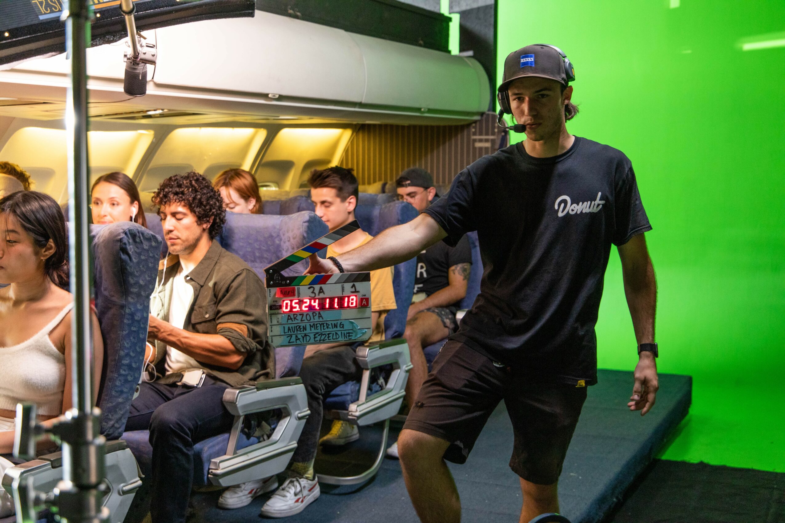 Man in black shirt and cap standing on a green screen set, with seated passengers behind him, some looking at the camera.