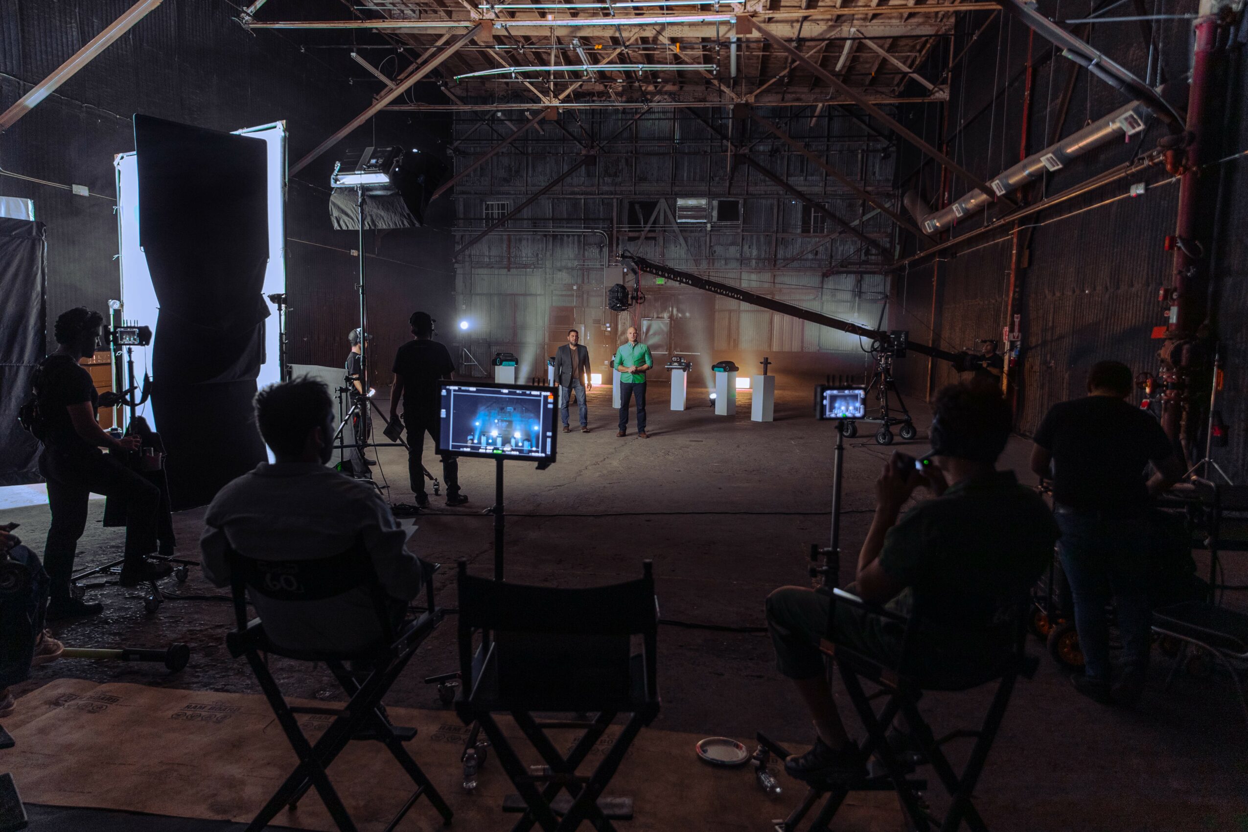 People in a studio or stage setting with lighting and equipment, some seated and some standing, facing a person speaking.