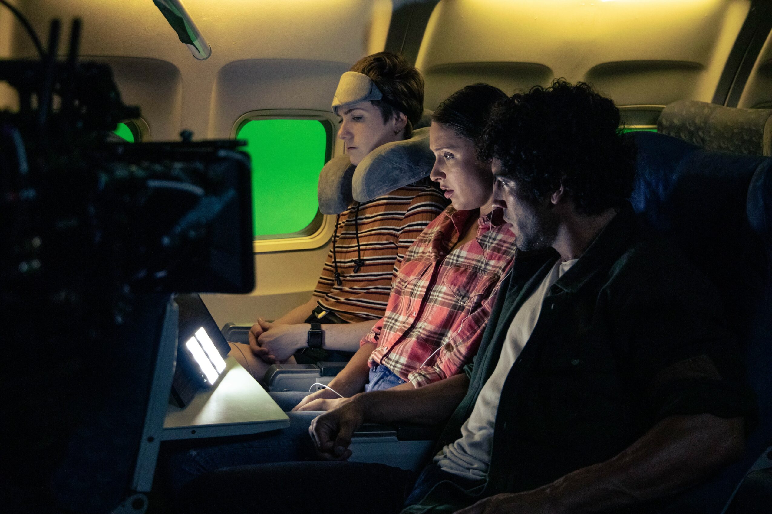Three people sitting in an airplane, looking at screens, with a green screen visible outside the window.