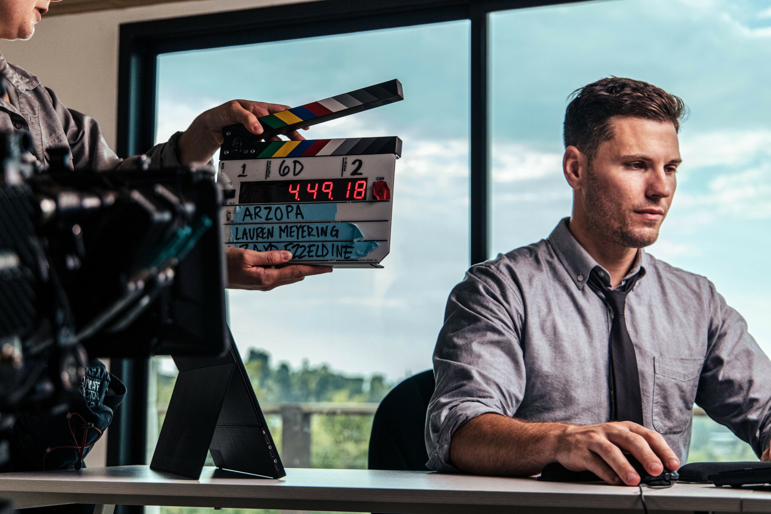 Man in gray shirt working at desk with a window showing trees and sky behind him.