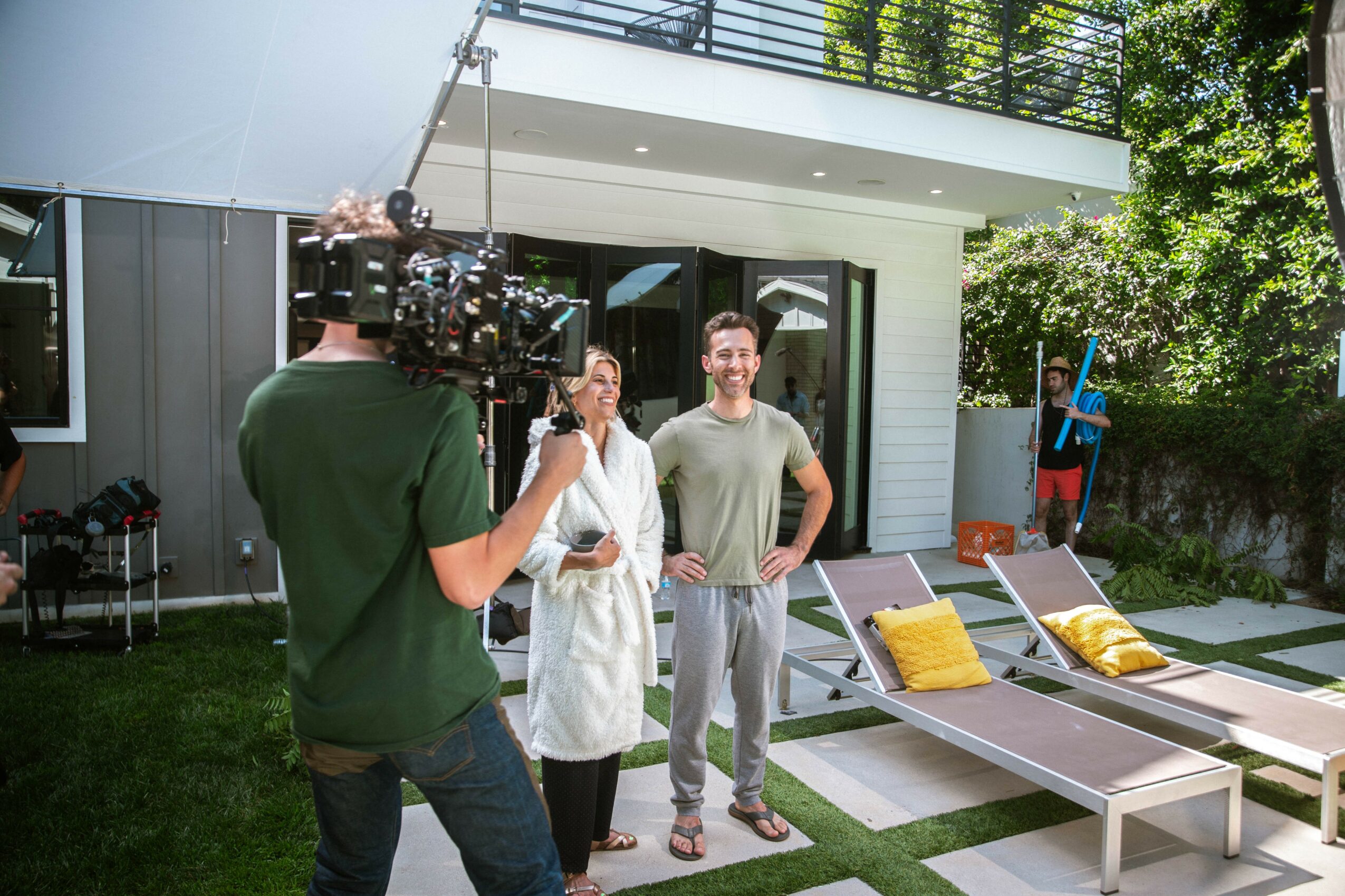 Two people being filmed outdoors near lounge chairs, with a camera operator recording them, in a backyard setting.