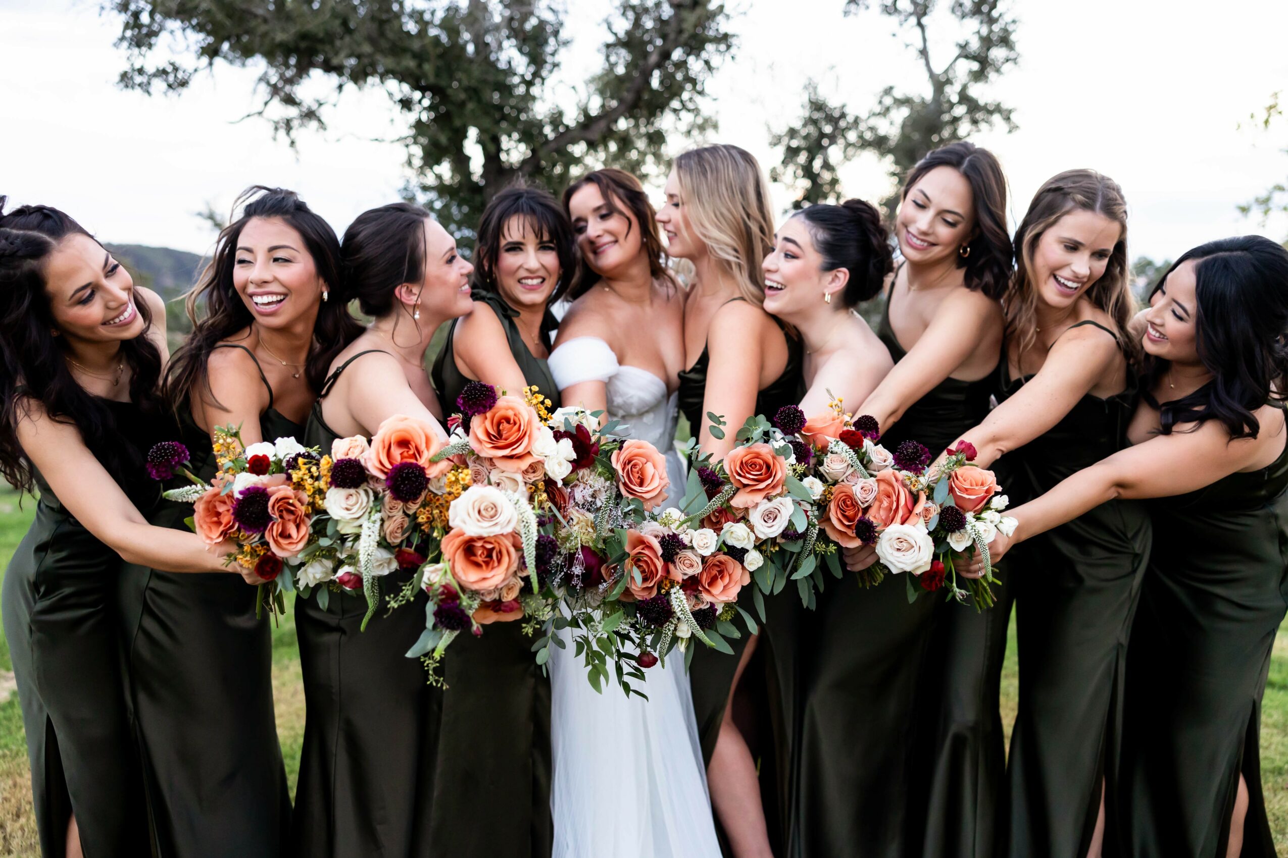 Group of women in black dresses holding a large floral arrangement outdoors, smiling and posing together.