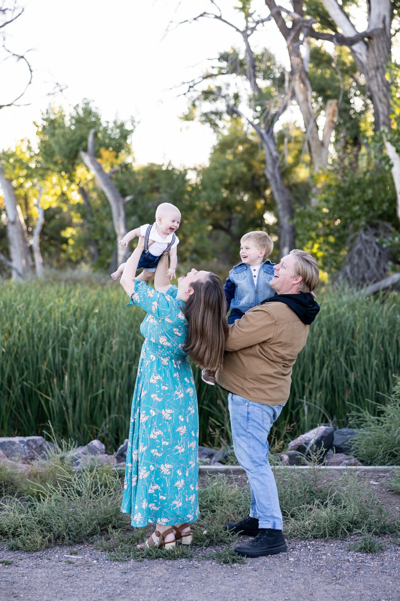 Family outdoors with trees and grass, a woman lifts a baby, a man holds a young boy, all smiling.