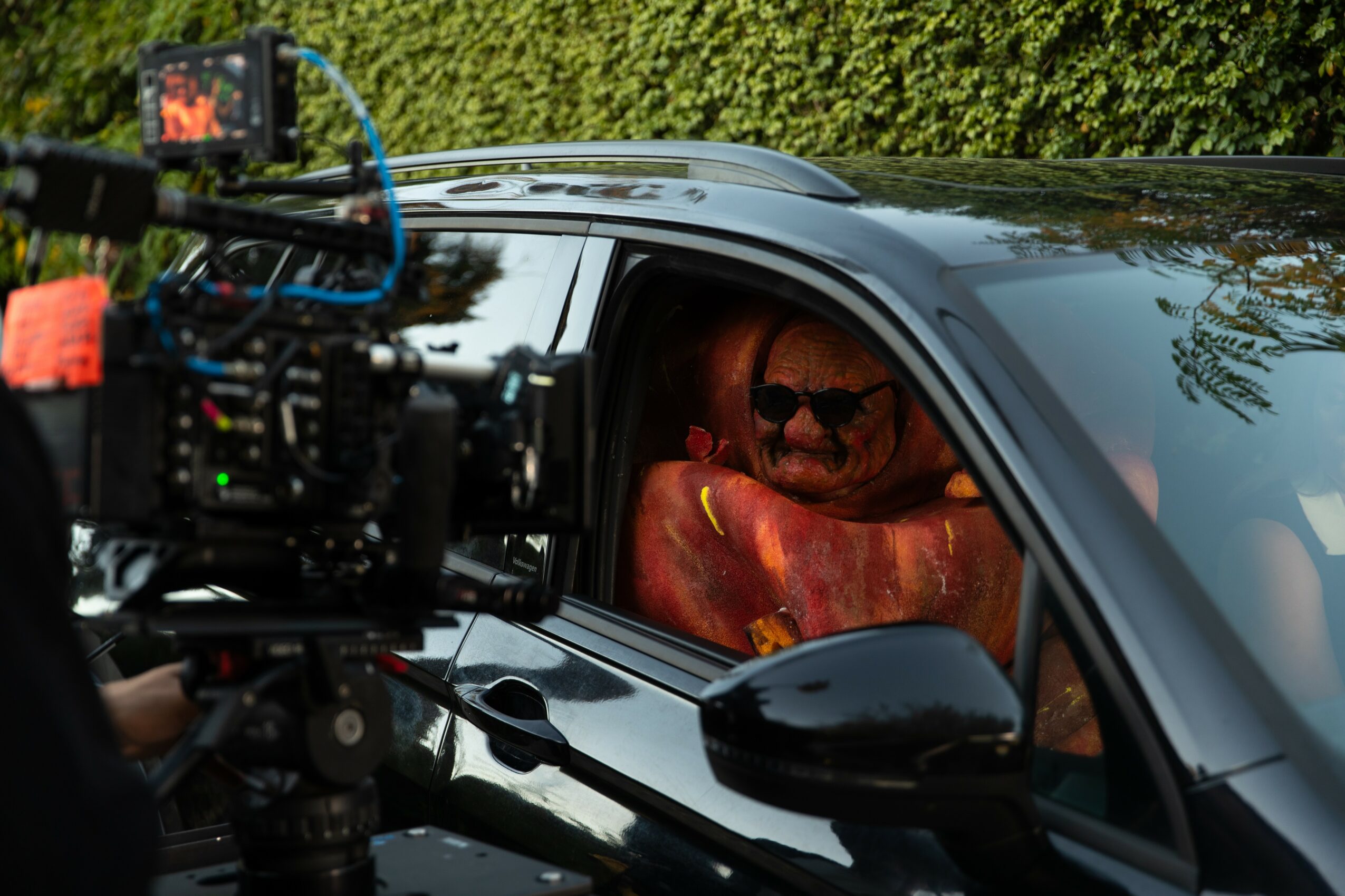 Person inside a car, visible through the window, with a camera filming the scene outside, greenery in the background.