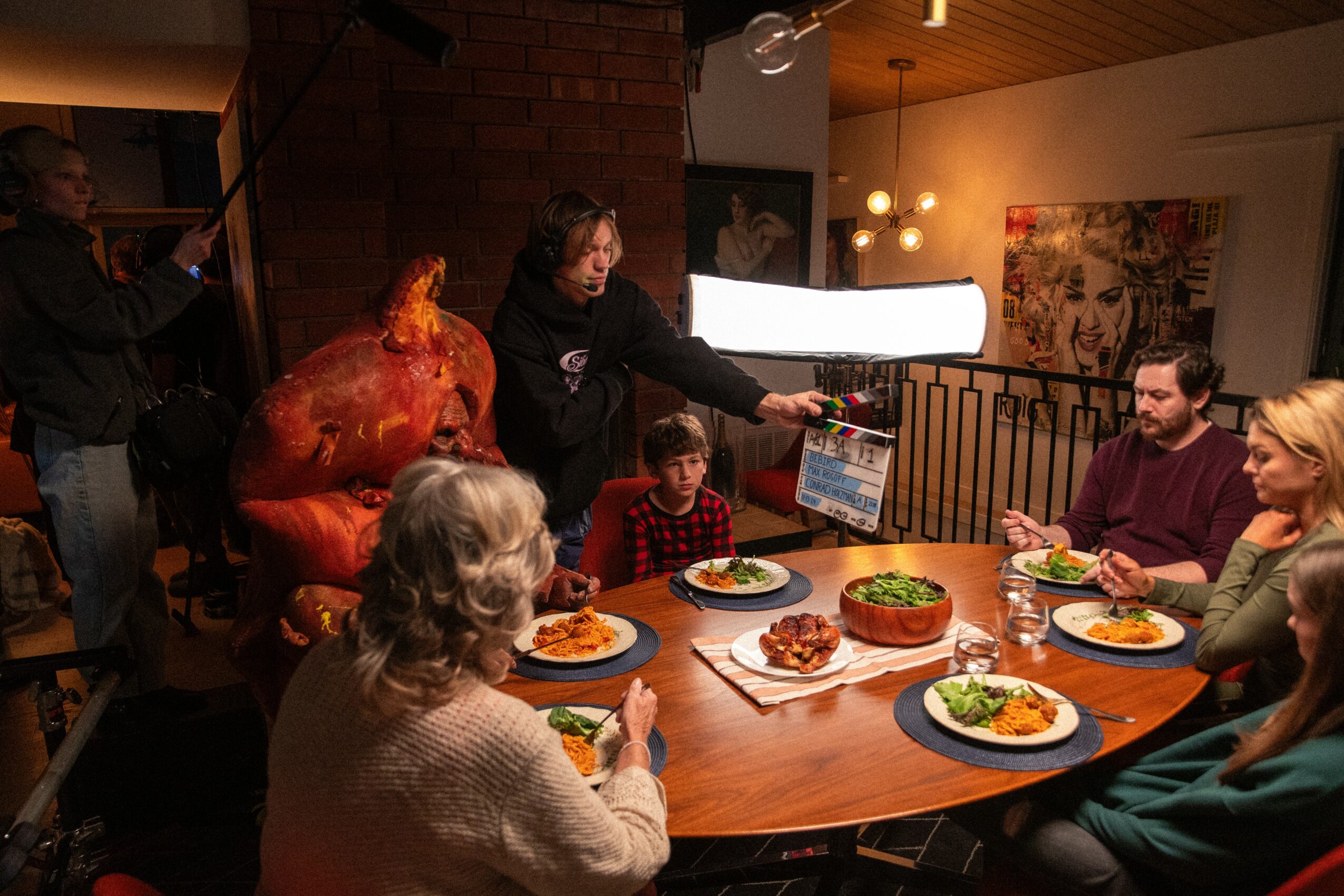 People gathered around a table with a cake, plates of food, and a person holding a book, in a cozy room.