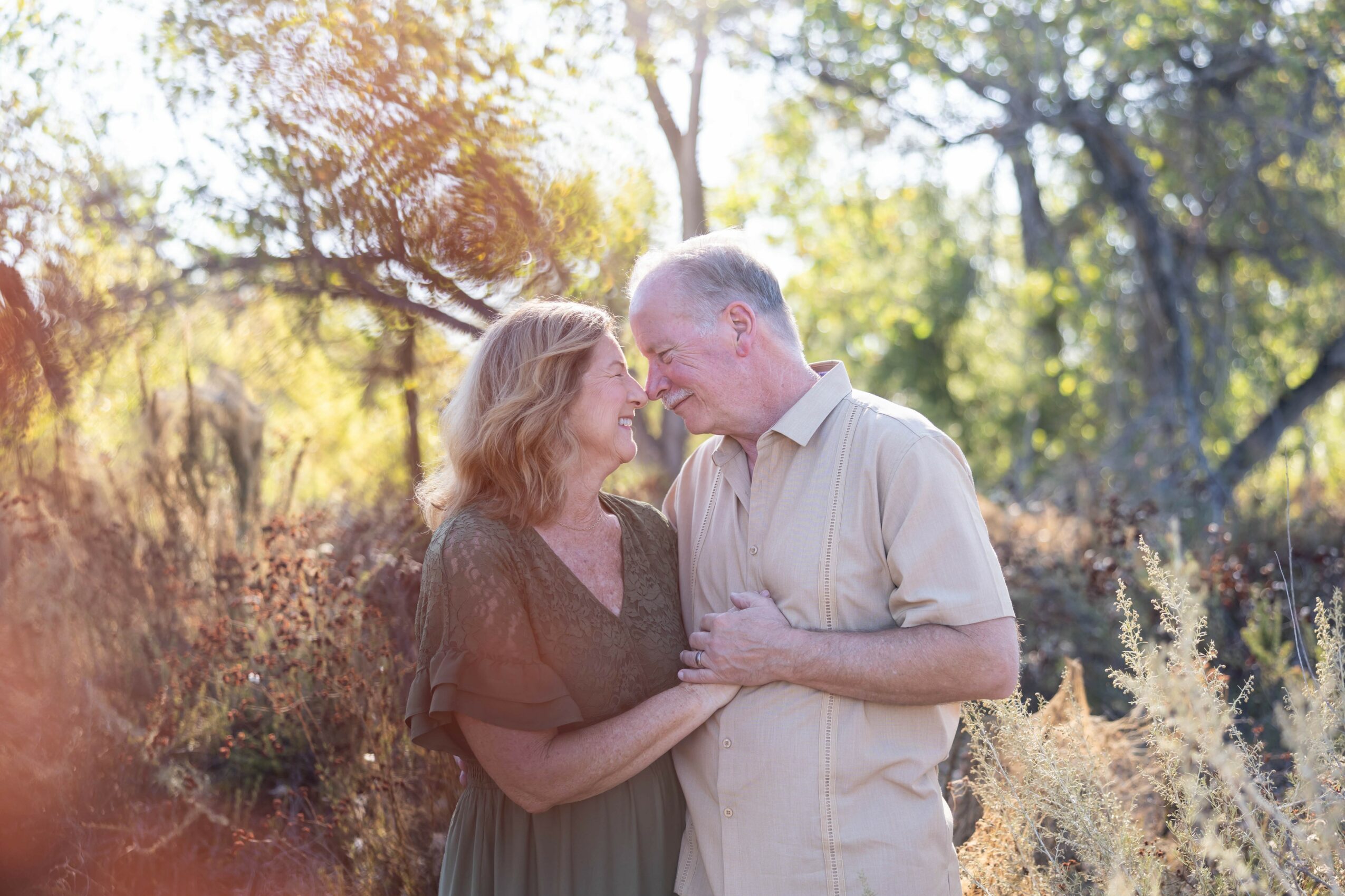 A couple stands close together outdoors, smiling and touching foreheads, surrounded by trees and sunlight.
