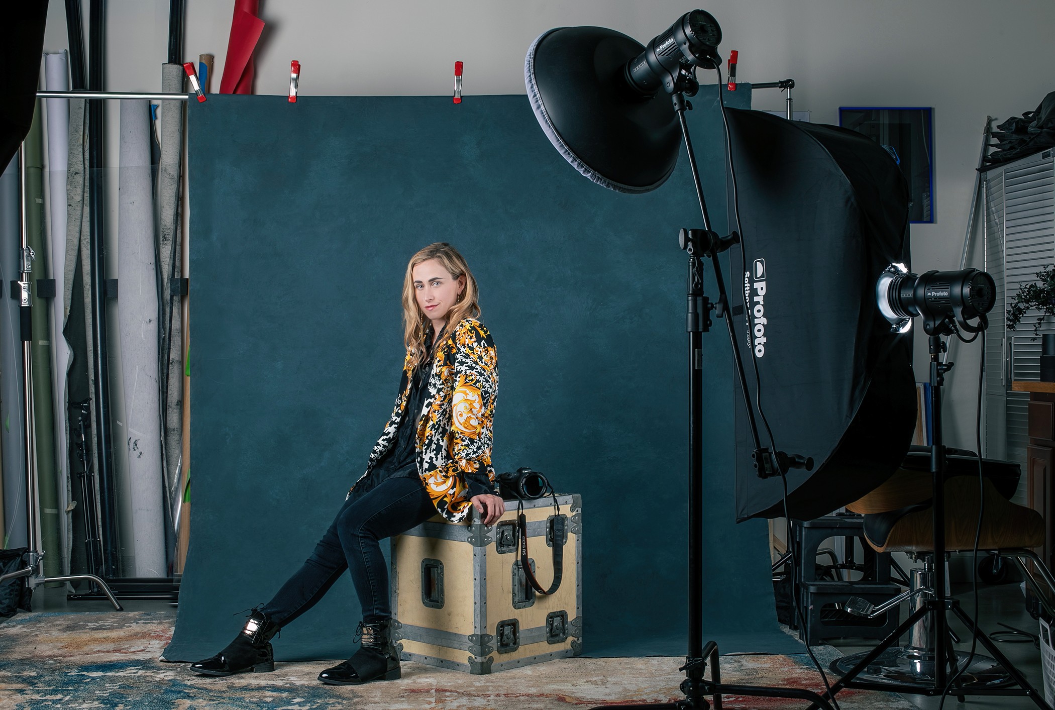 Young woman sitting on a large suitcase in a photography studio with equipment and a dark green backdrop.