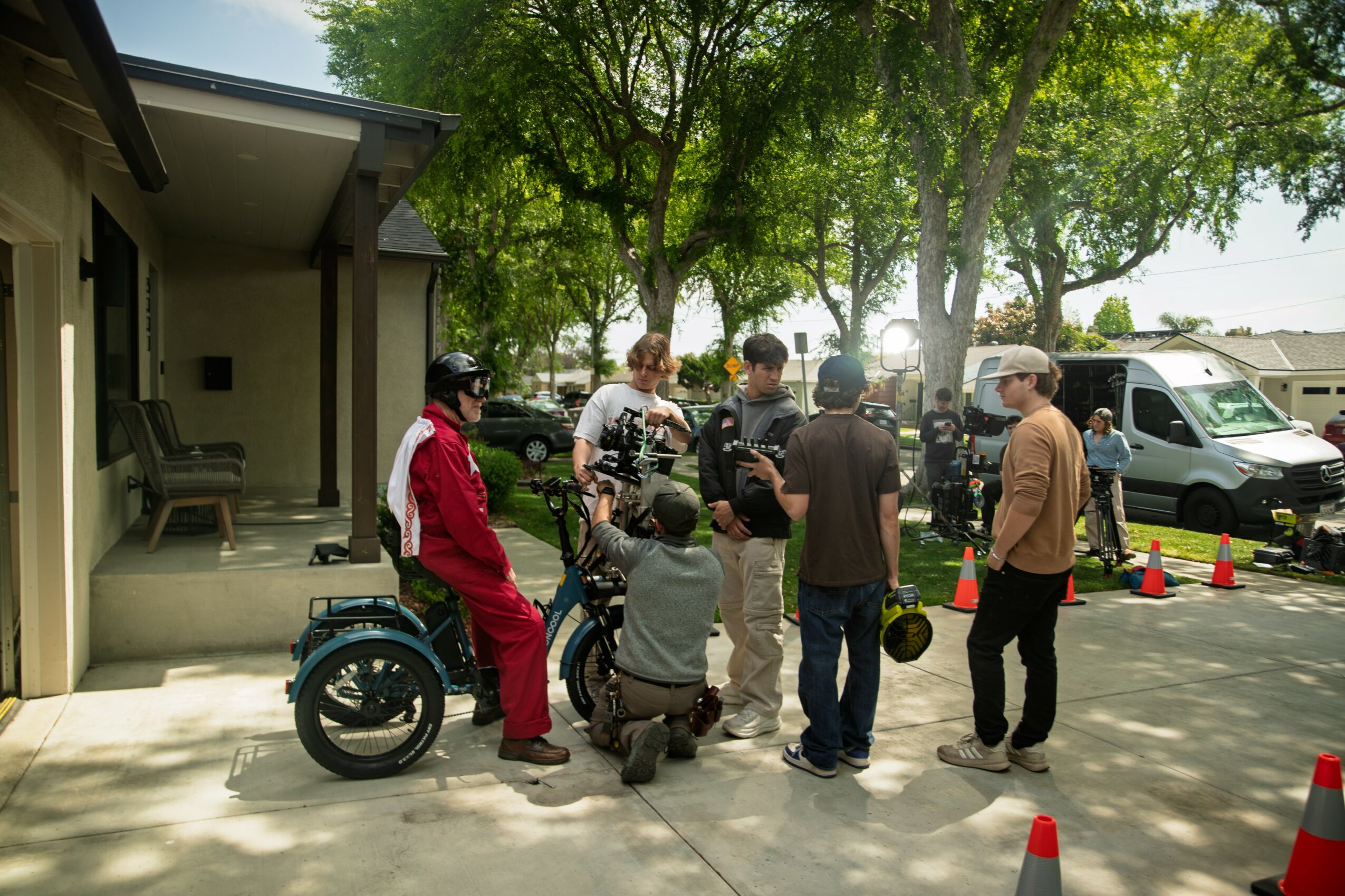 Group of people gathered outdoors near a motorcycle, with trees and parked vehicles in the background.