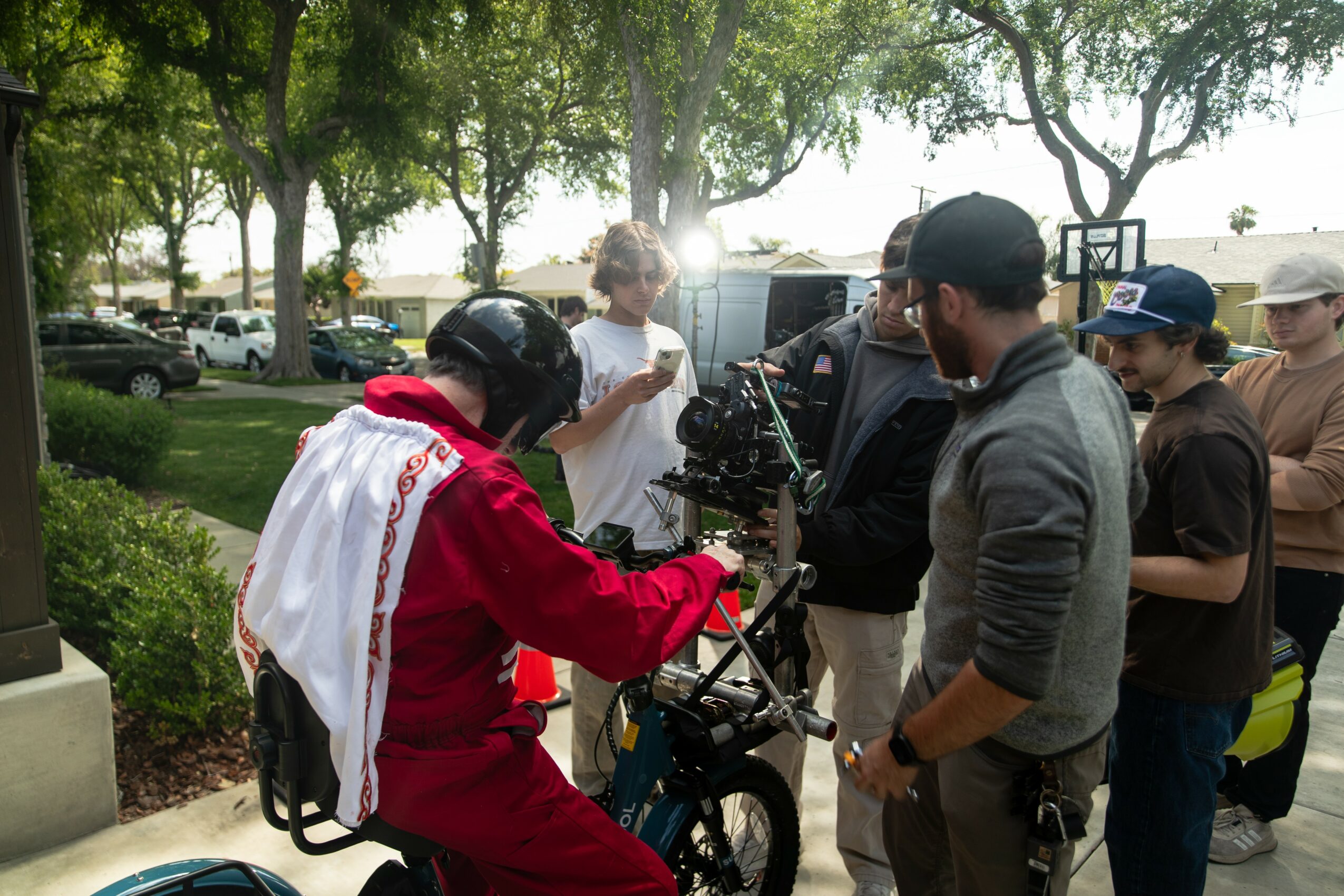 Group of people gathered outdoors near trees, some wearing helmets, with a person on a bicycle and others talking.