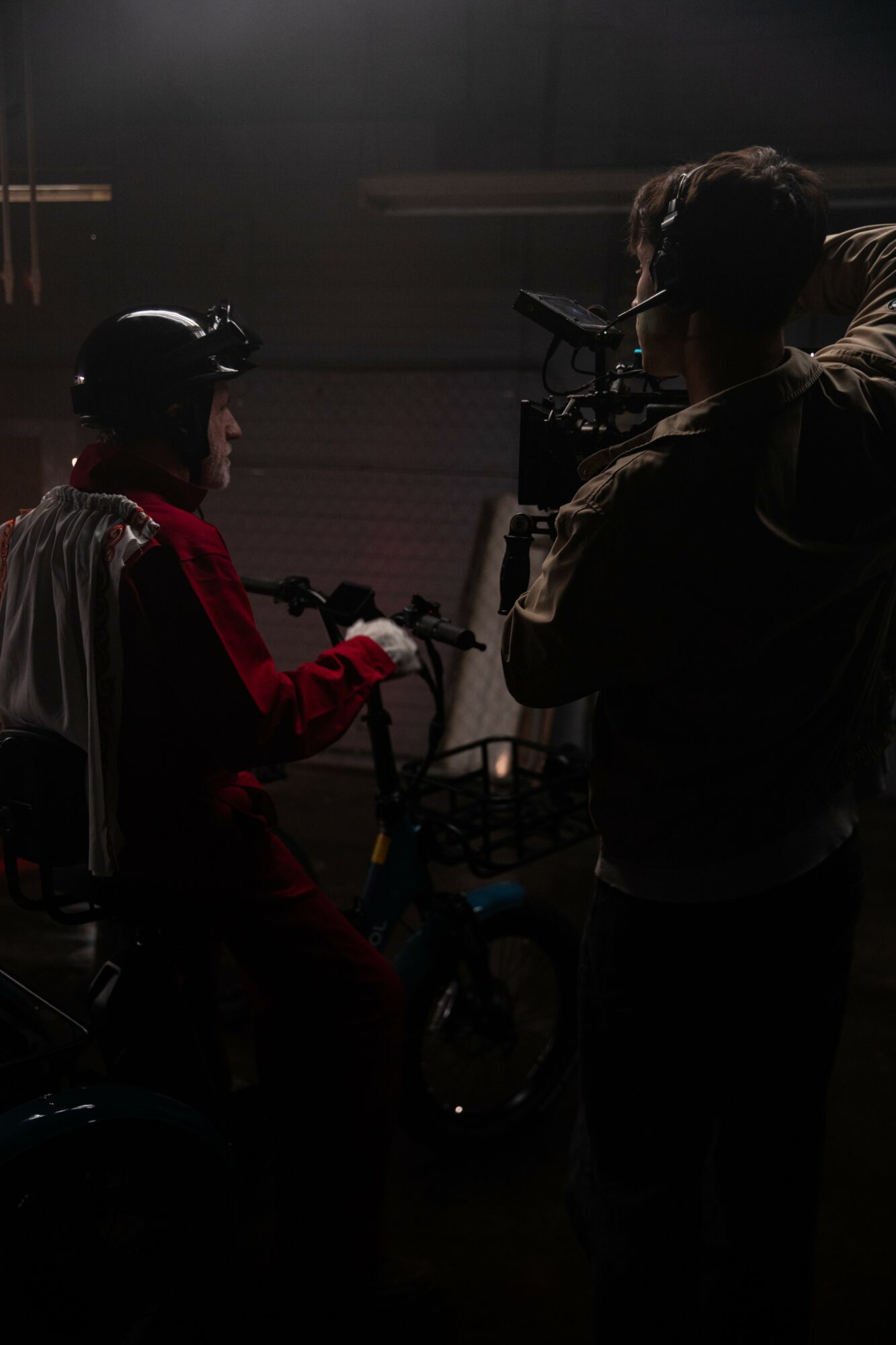 Person in red jacket with helmet on bicycle, being filmed by a cameraman, in a dimly lit setting.