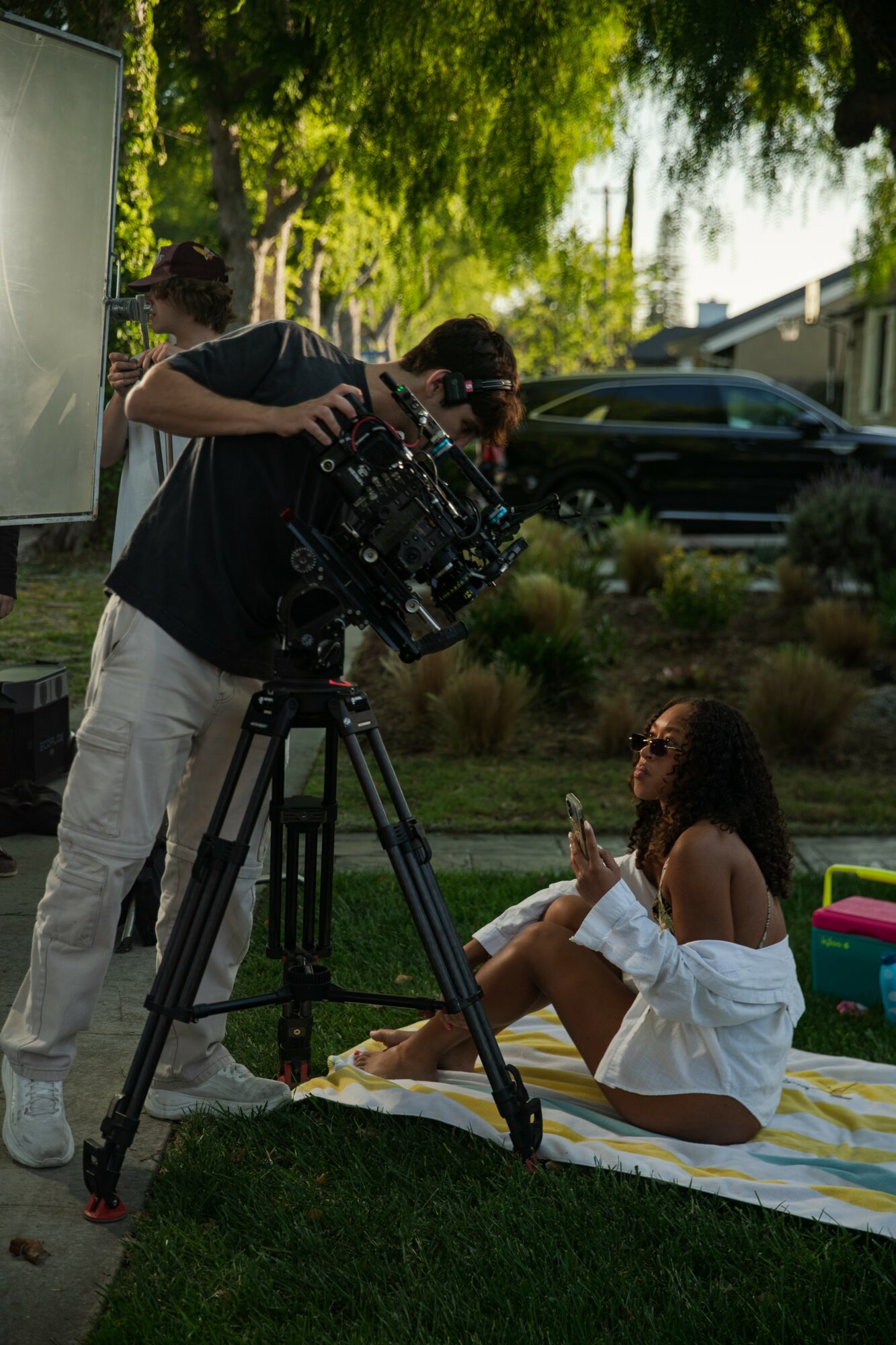 Person operating camera on tripod filming a woman sitting on a blanket outdoors, trees and cars in background.
