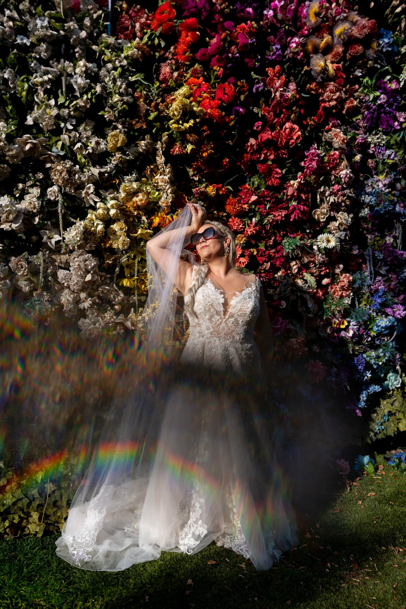 Bride in a white wedding dress and veil stands in front of a colorful flower wall, shielding her eyes from sunlight.