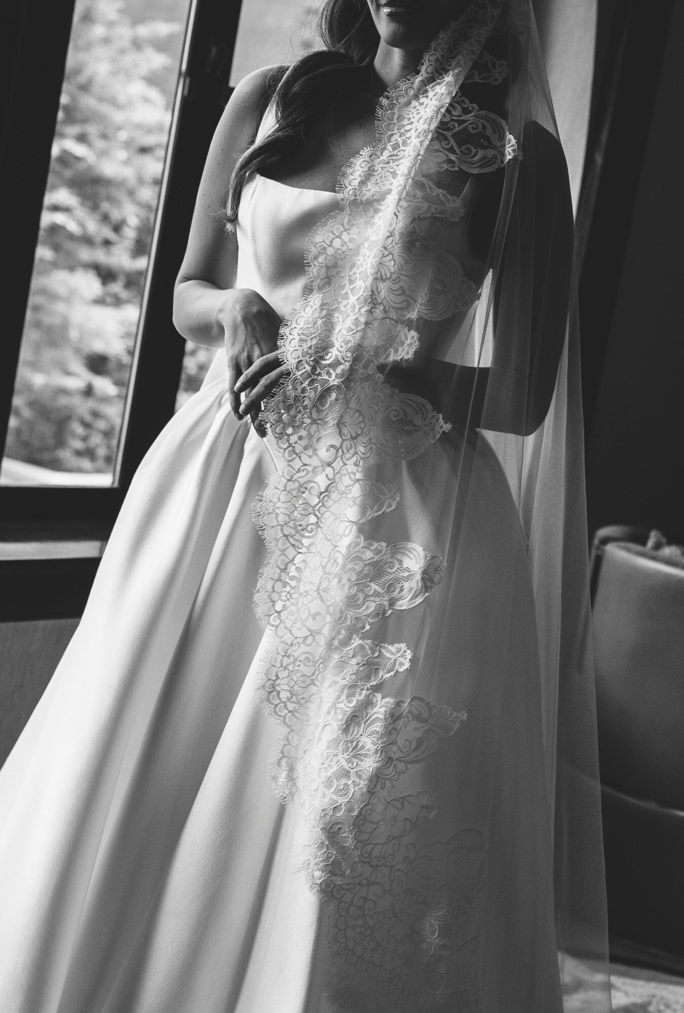 Woman in wedding dress standing near window with lace veil, natural light, indoor setting.
