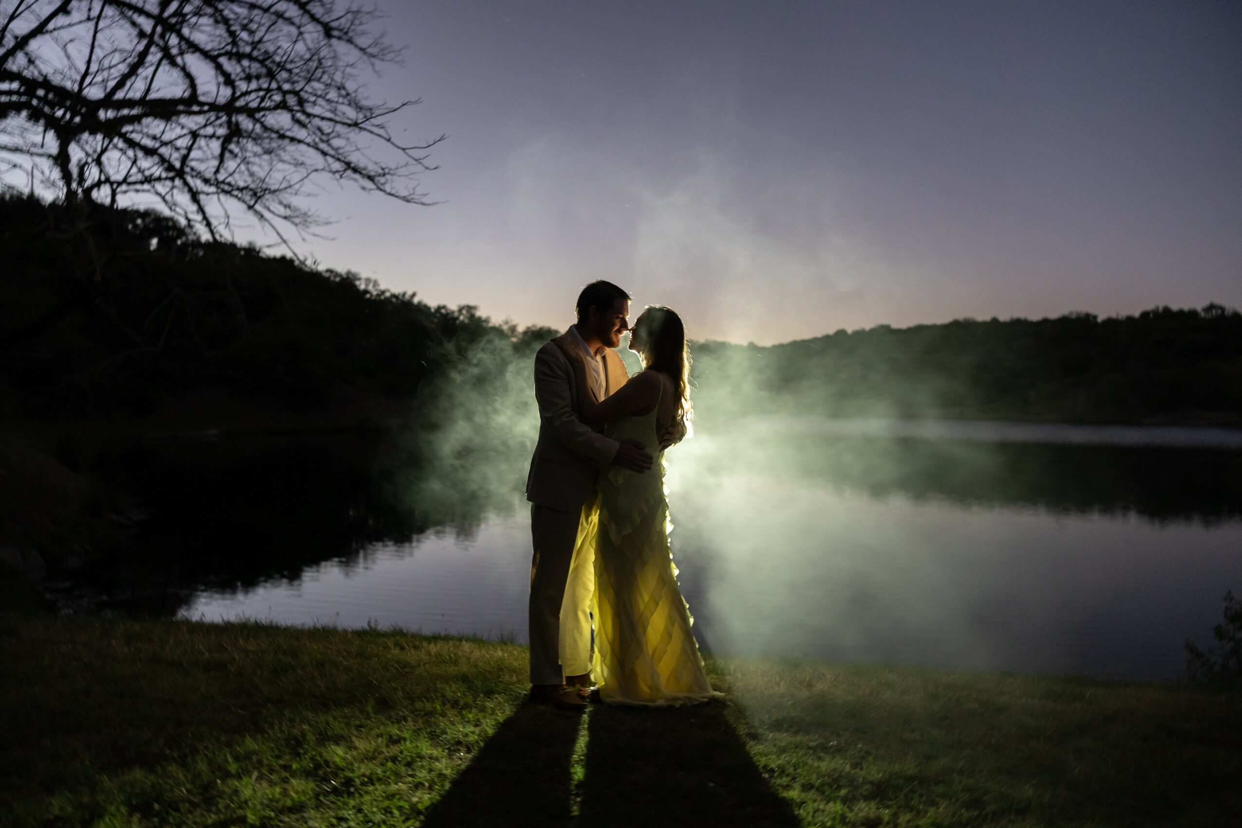 Silhouette of a couple embracing by a lake at sunset, with trees and hills in the background.