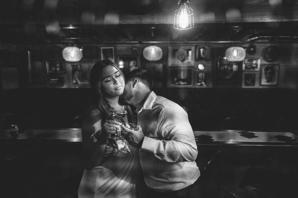Two people sit close together in a dimly lit setting, looking at a phone, with hanging lights and framed pictures in the background.