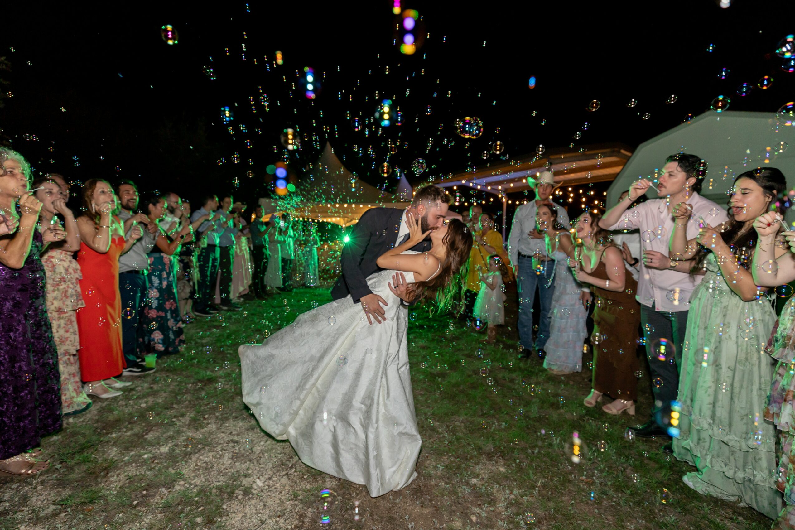 Couple dancing in the rain with guests clapping and celebrating around them at night, colorful lights in the background.