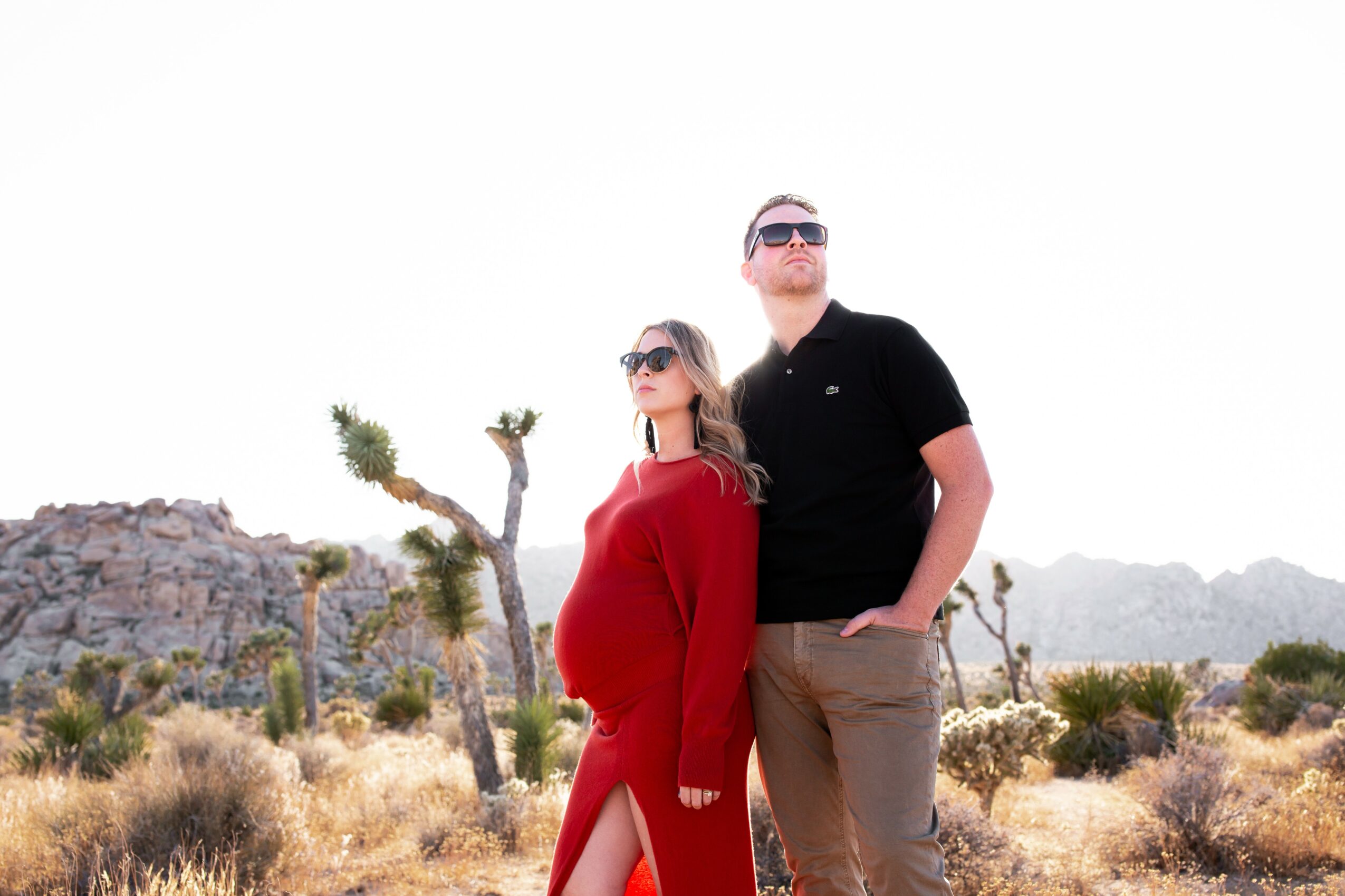 A pregnant woman and a man stand outdoors in a desert landscape with cacti and rocky hills, both wearing sunglasses.