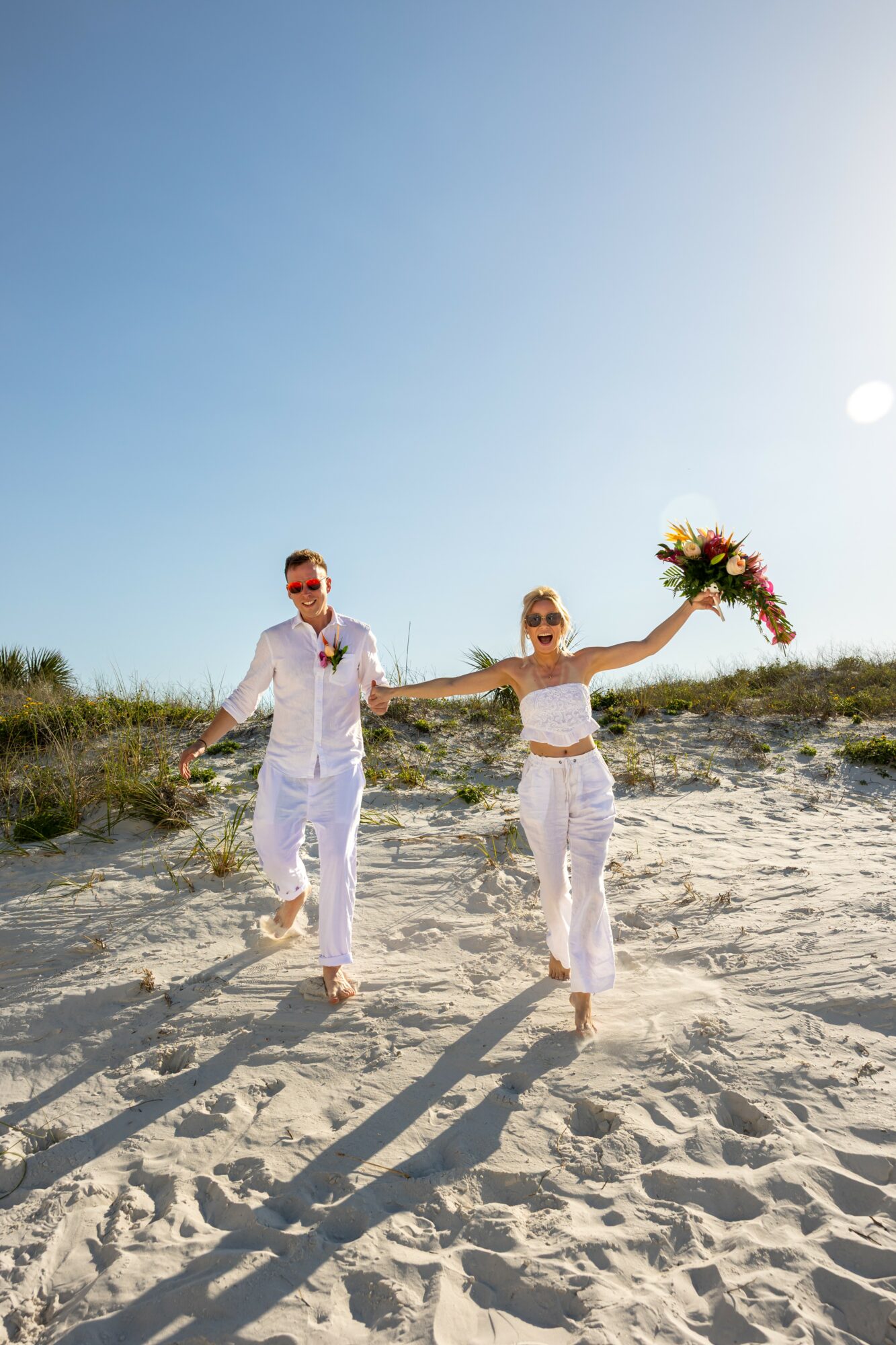 Two people walk on sandy beach holding hands, one holding a bouquet of flowers, with clear blue sky above.