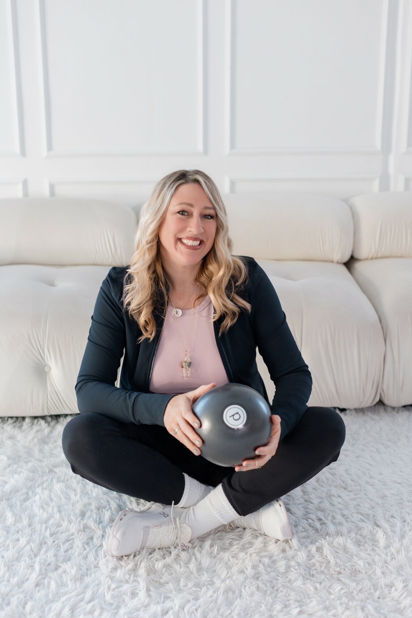 Woman sitting cross-legged on a white carpet, holding a black round object, smiling, in a bright room with white walls and sofa.