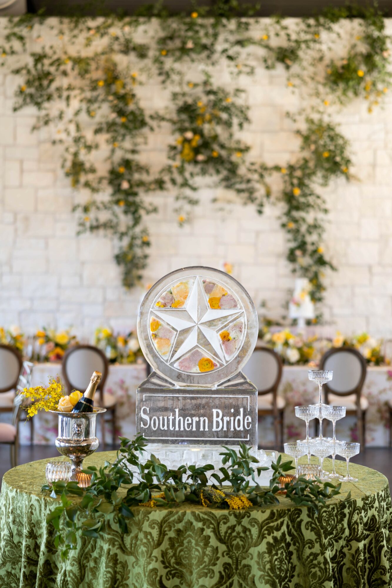 Table with a green patterned tablecloth, floral centerpiece, and chairs in front of a white brick wall with climbing plants.