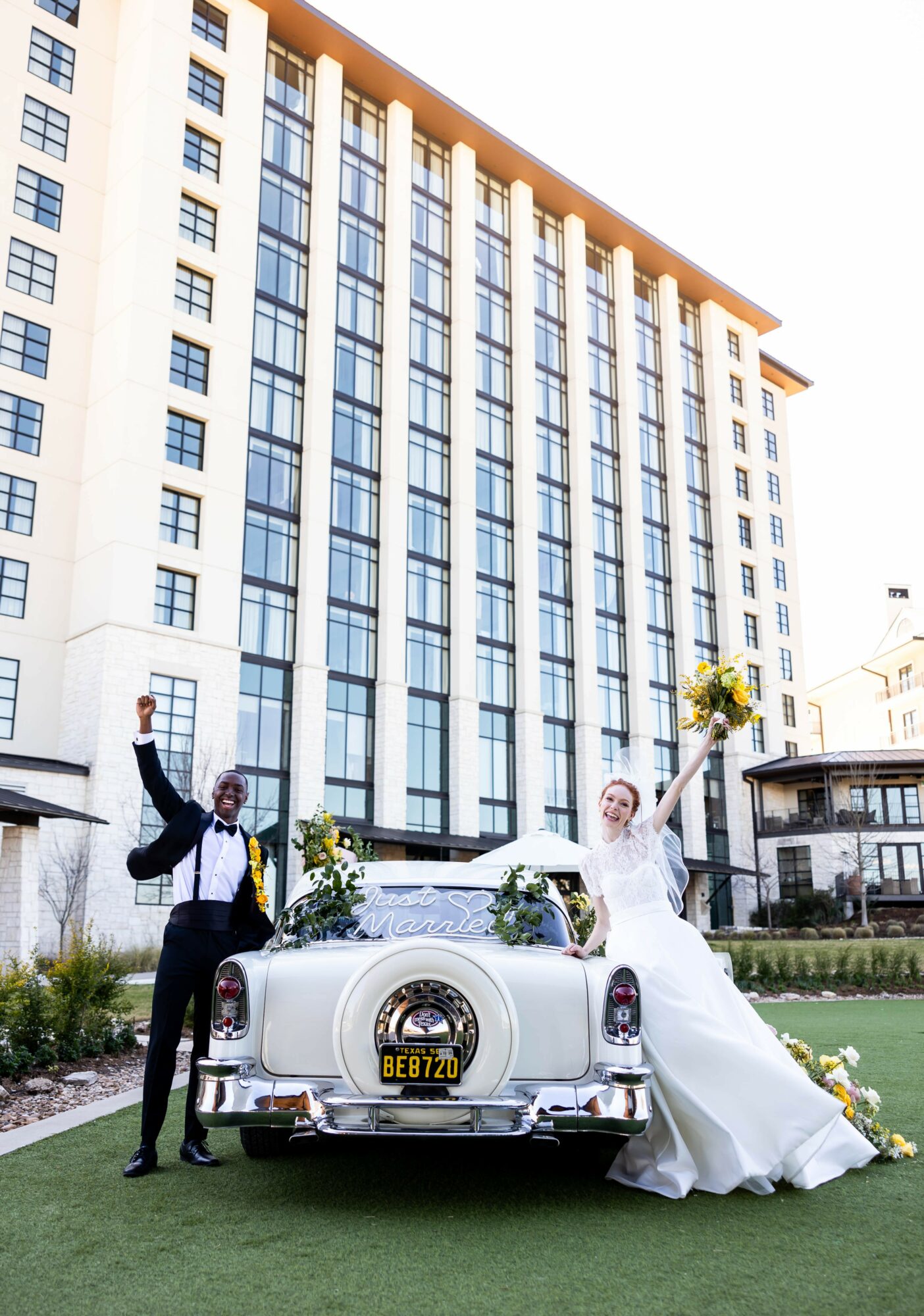 Bride and groom celebrating next to a vintage car in front of a tall building, holding flowers and smiling.
