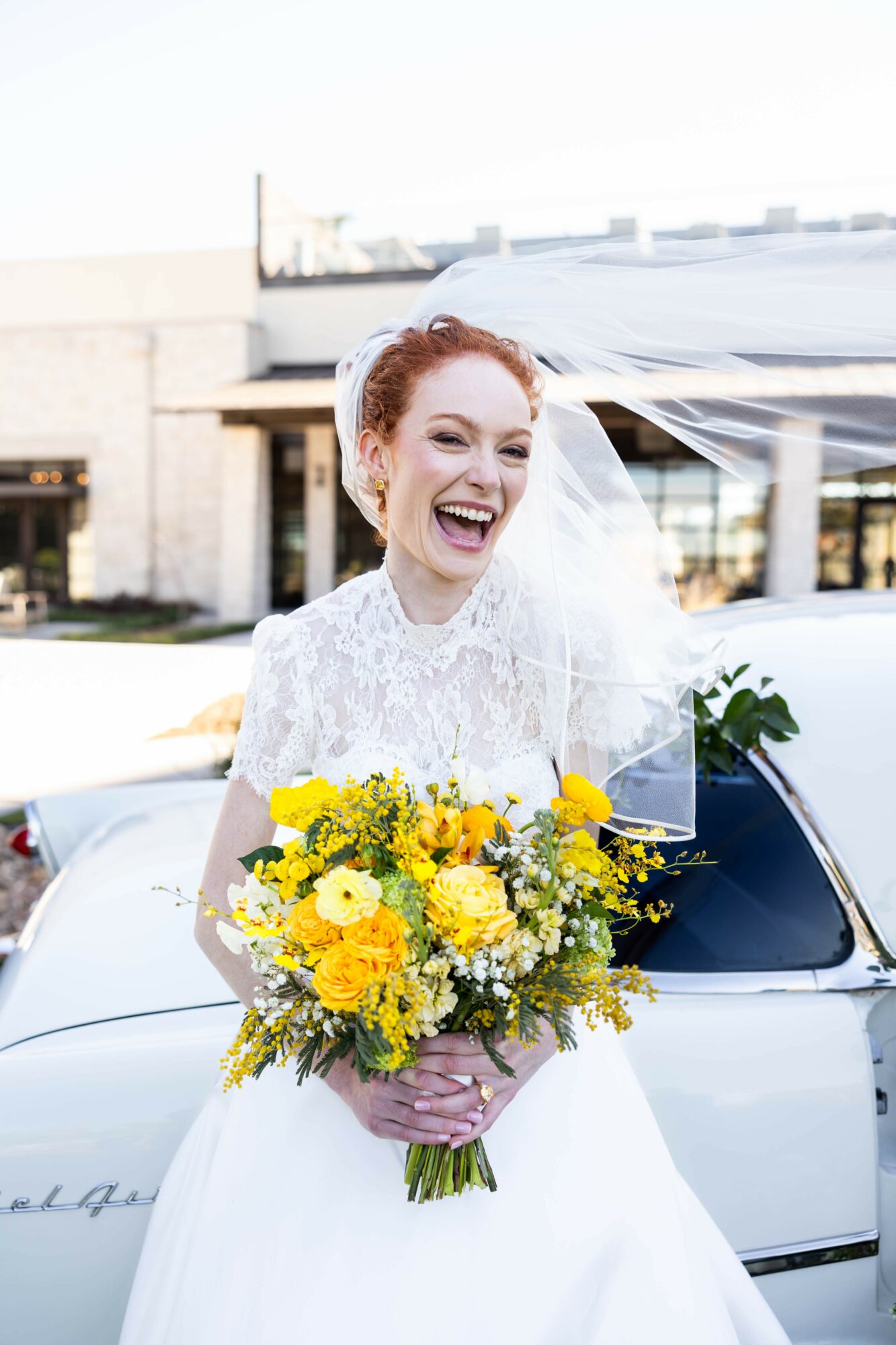 Smiling bride with red hair holding a yellow flower bouquet outdoors, wearing a white wedding dress and veil.