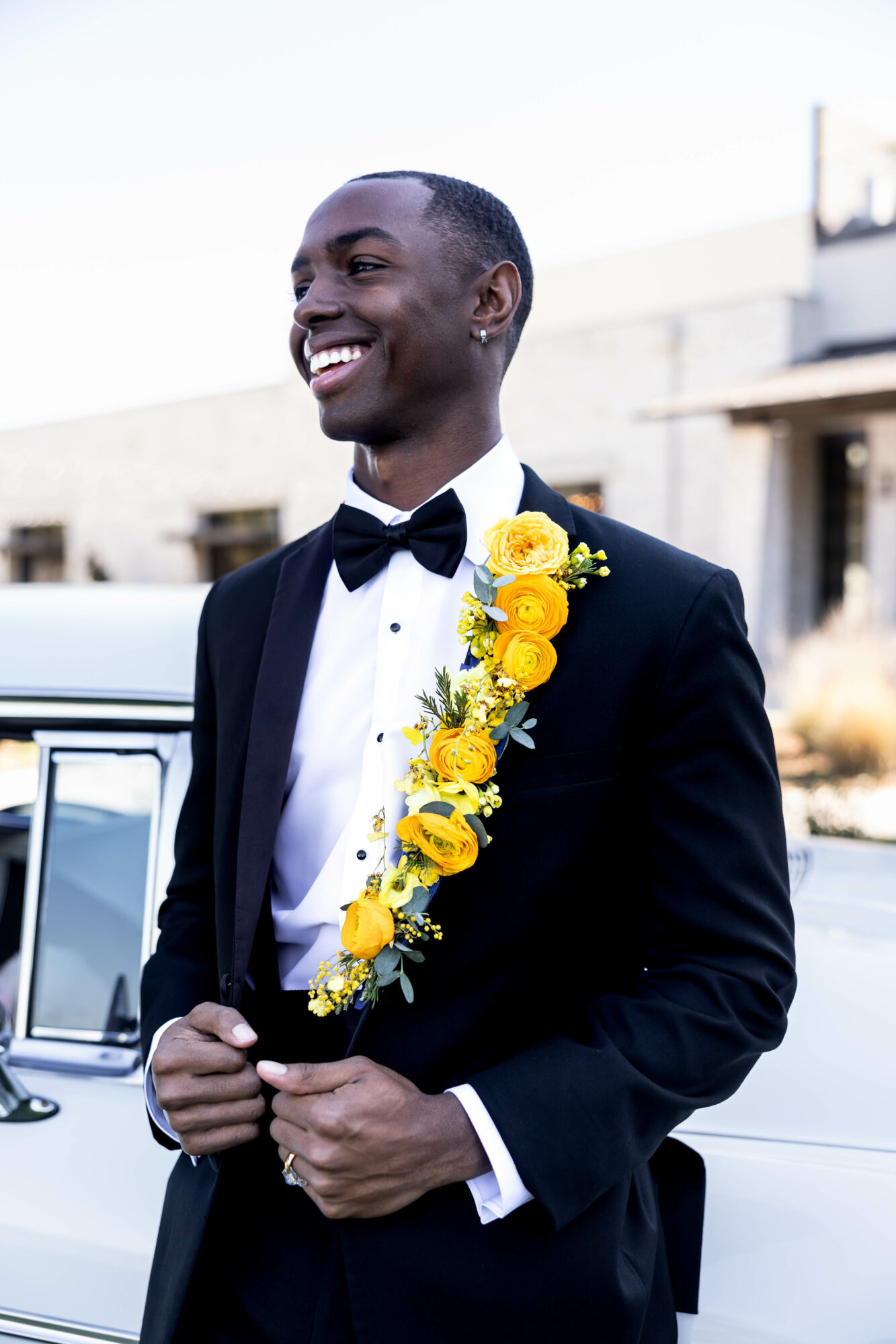 Man in tuxedo with a yellow floral boutonniere, smiling outdoors, with buildings and a car in the background.