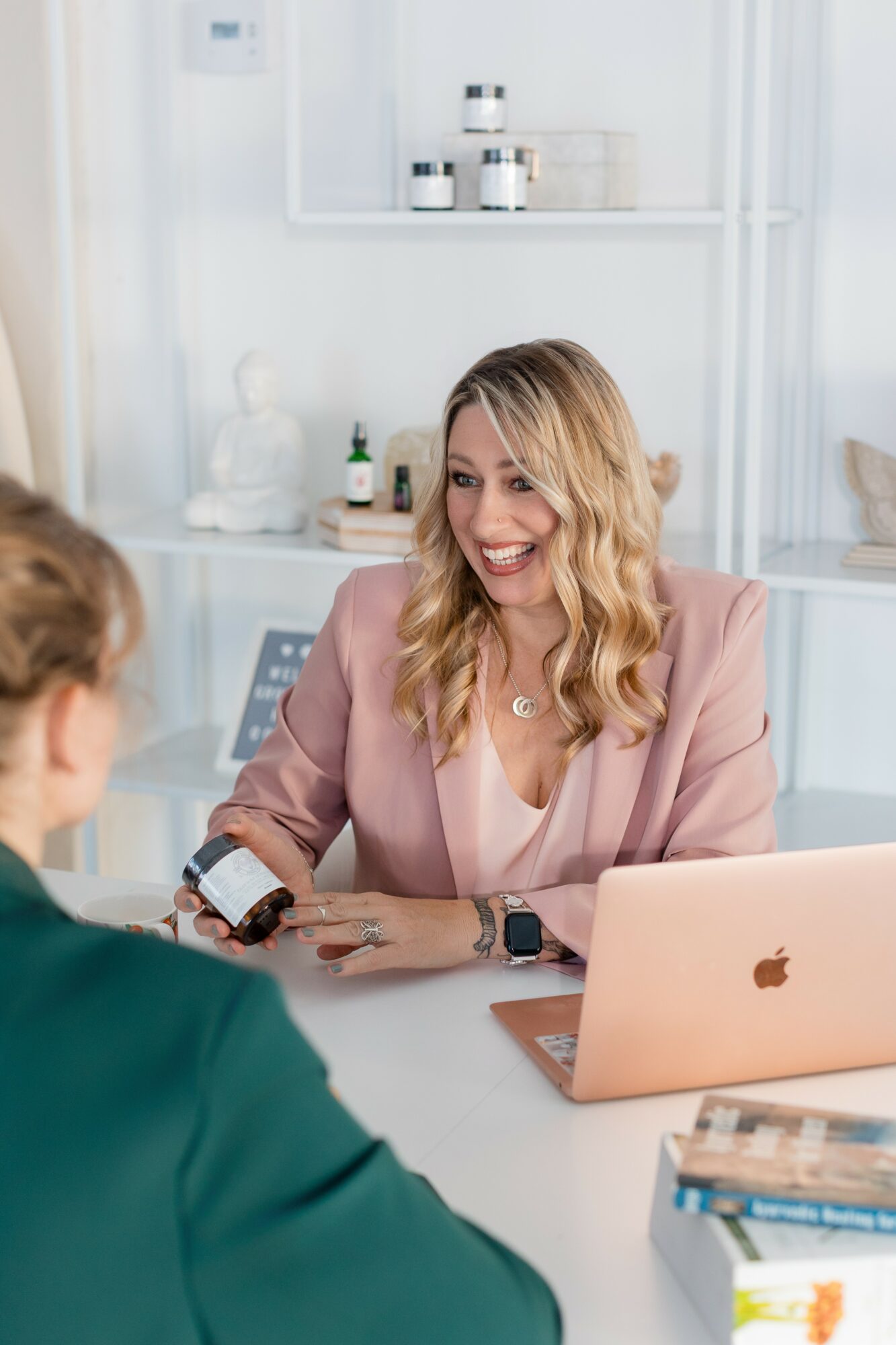 Woman with blonde hair smiling and talking to a person in a green shirt in a bright office, with a laptop and books on the table.