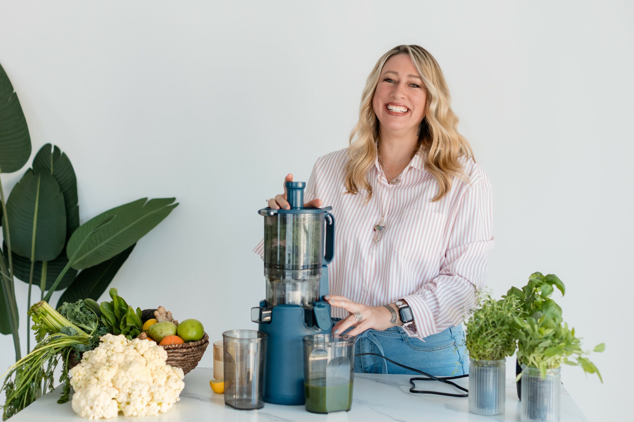 Woman smiling in a kitchen with fresh vegetables and a blender, wearing a striped shirt and jeans.