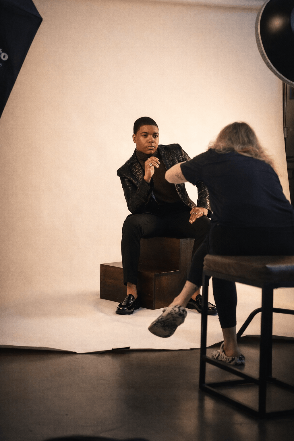 Person sitting on a stool, being photographed by a photographer with a backdrop, in a studio setting.