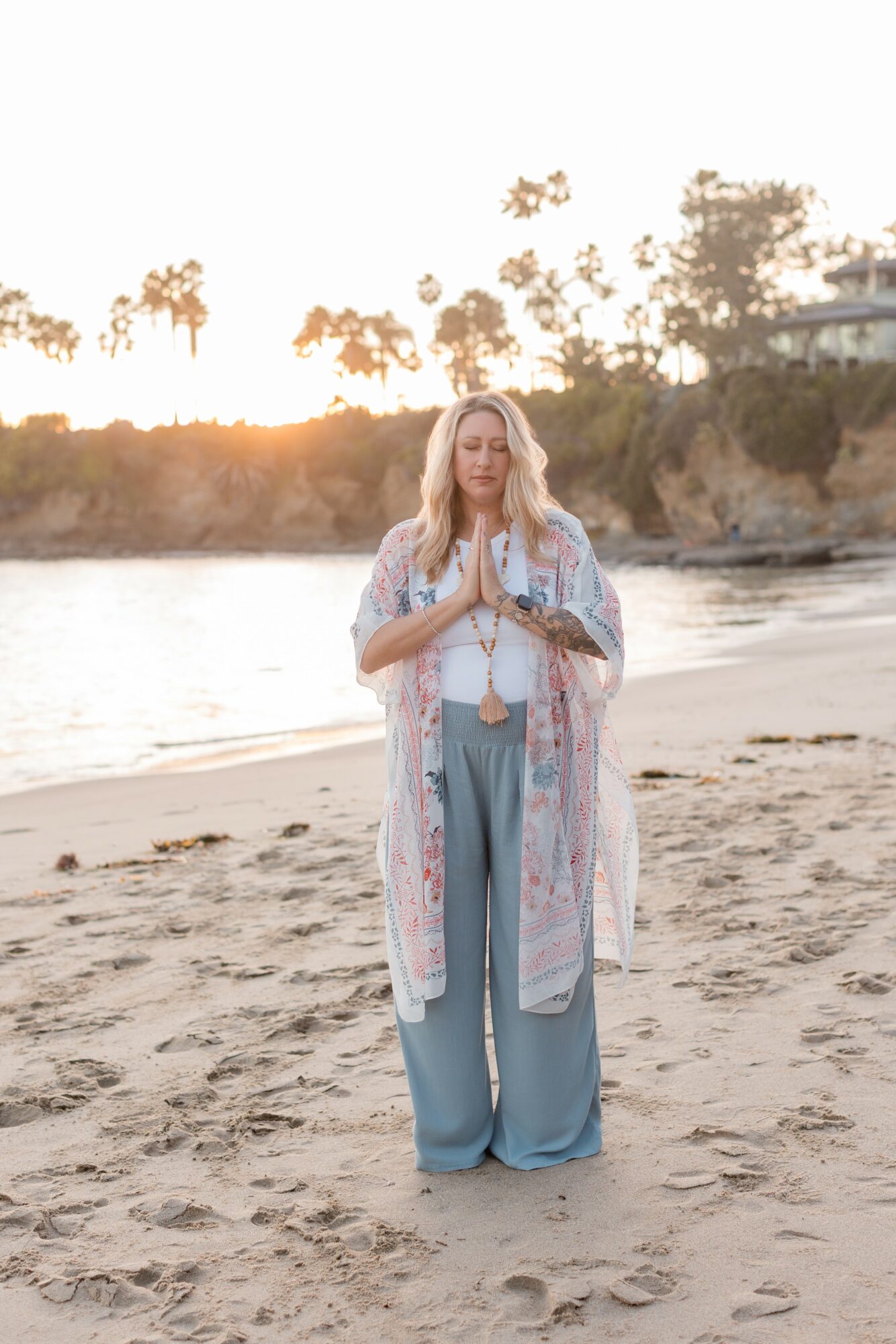Woman standing on beach with hands in prayer position, sunset in background, wearing flowy pants and a patterned cover-up.