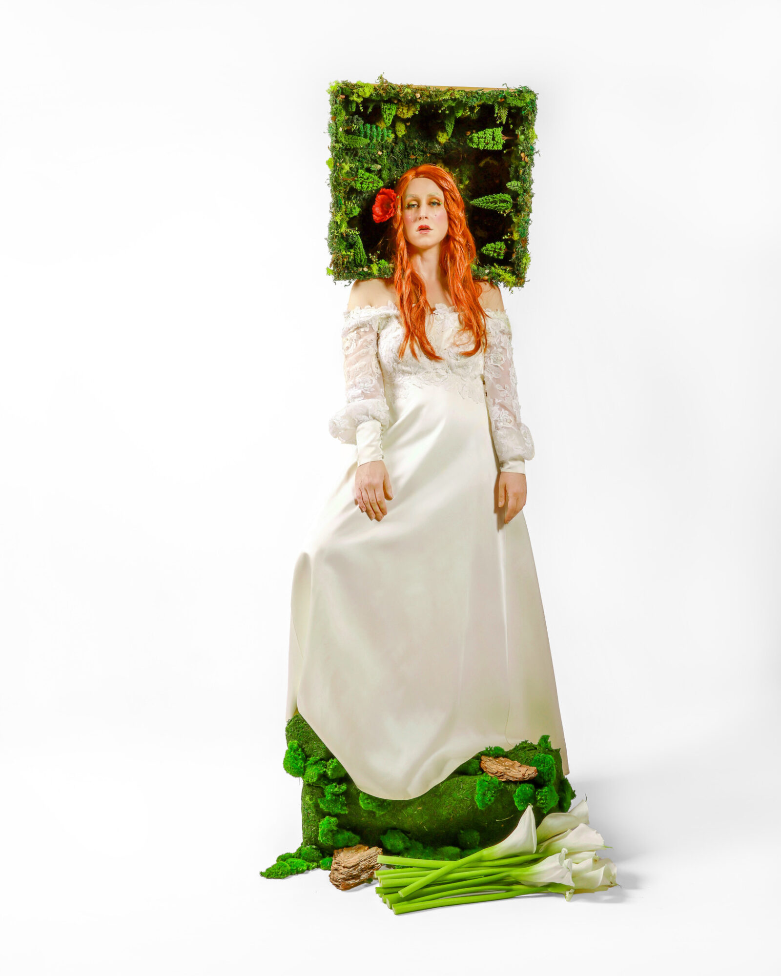 Woman with long red hair in a white dress standing in front of a green moss-covered frame, with plants at her feet.