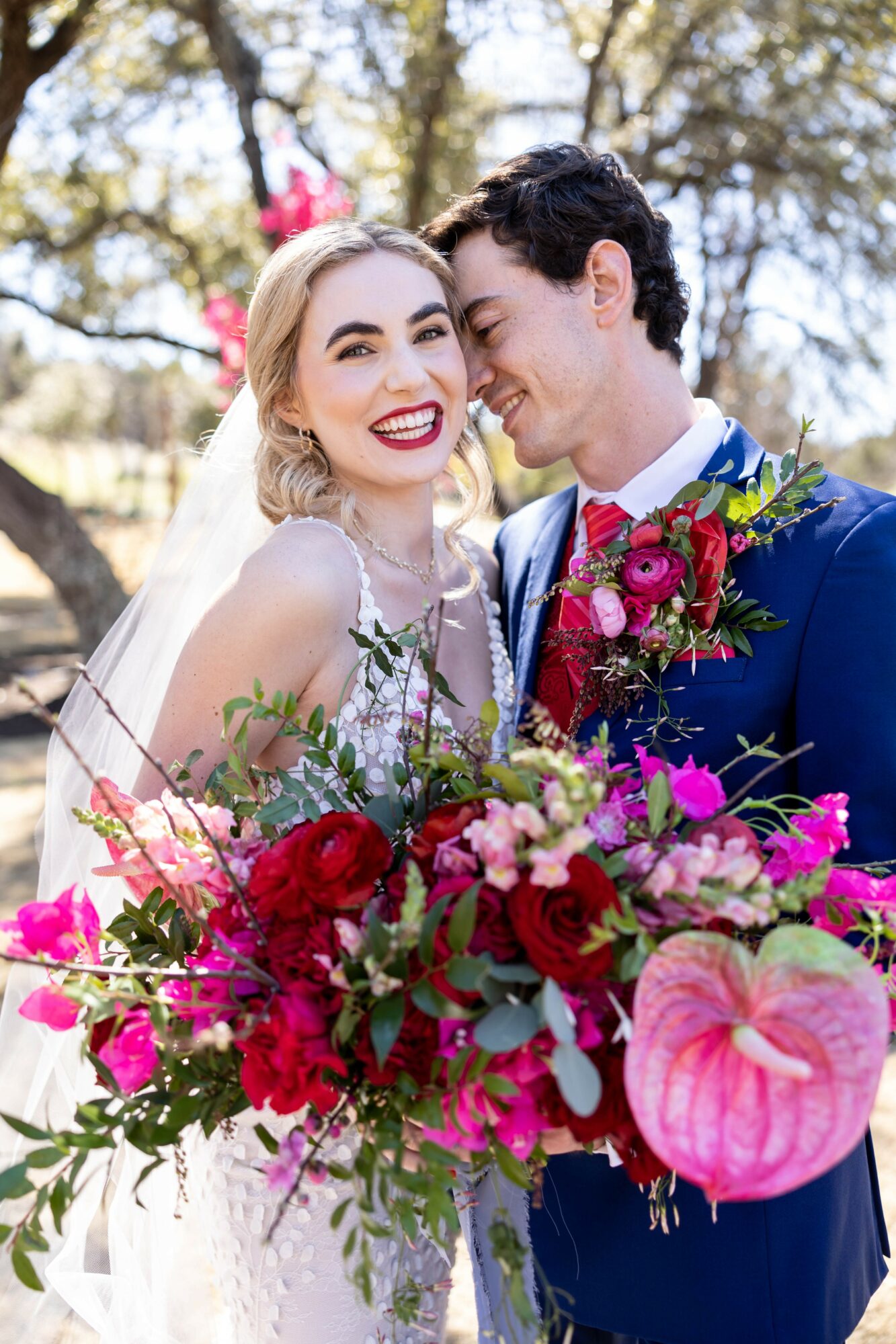 Bride and groom smiling outdoors, holding a large colorful bouquet of flowers, with trees in the background.