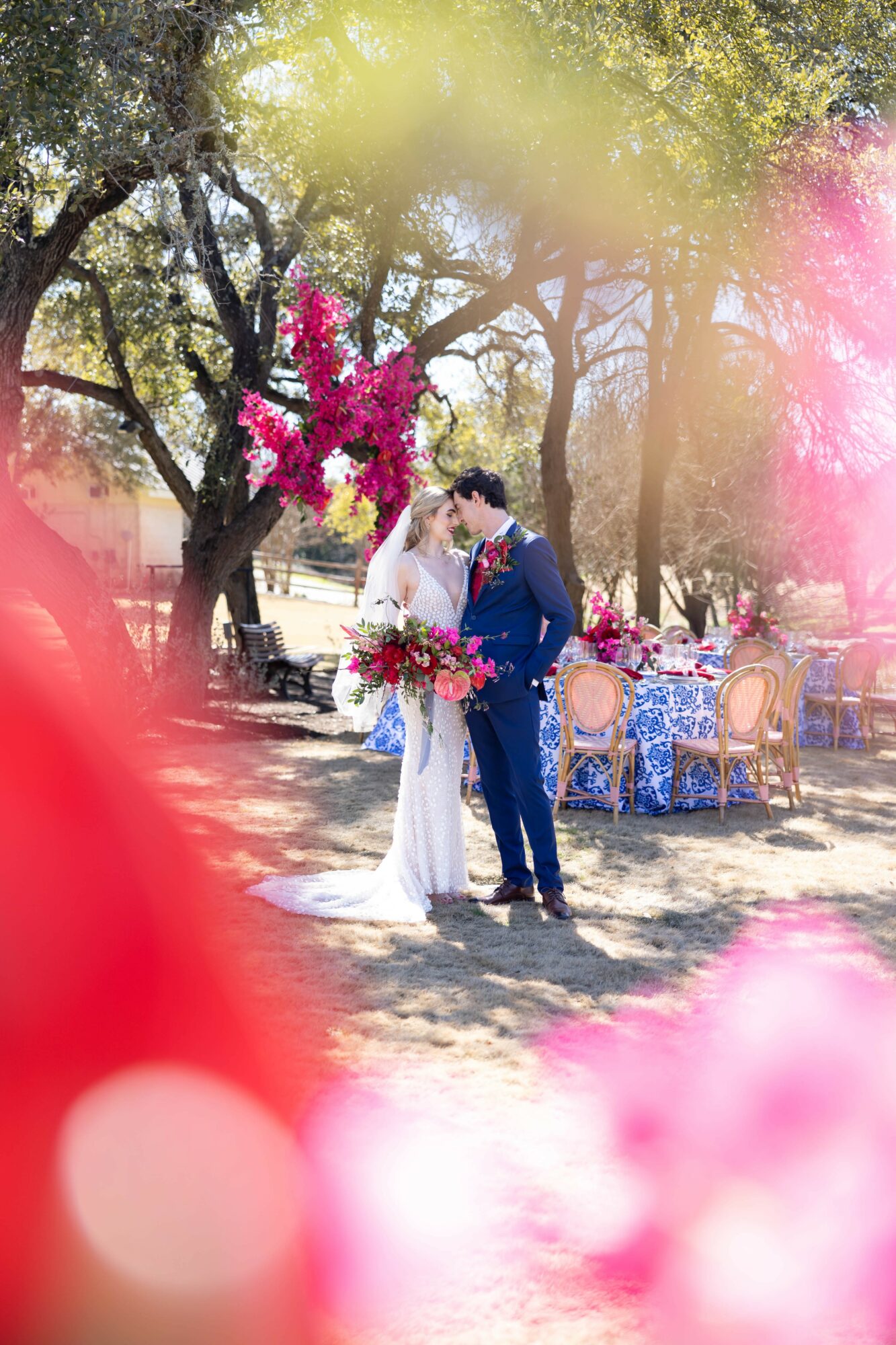 Bride and groom kissing outdoors with pink flowers and decorated tables in background.