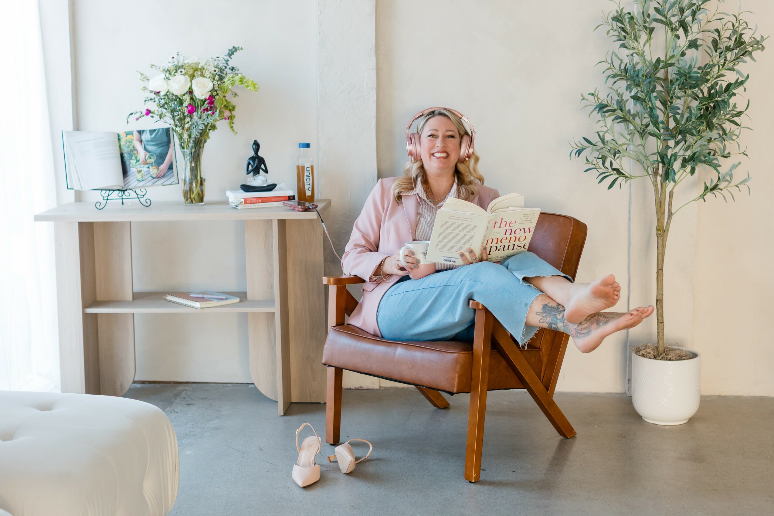 Woman sitting on a chair reading a book, smiling, with a plant and a table in the background.