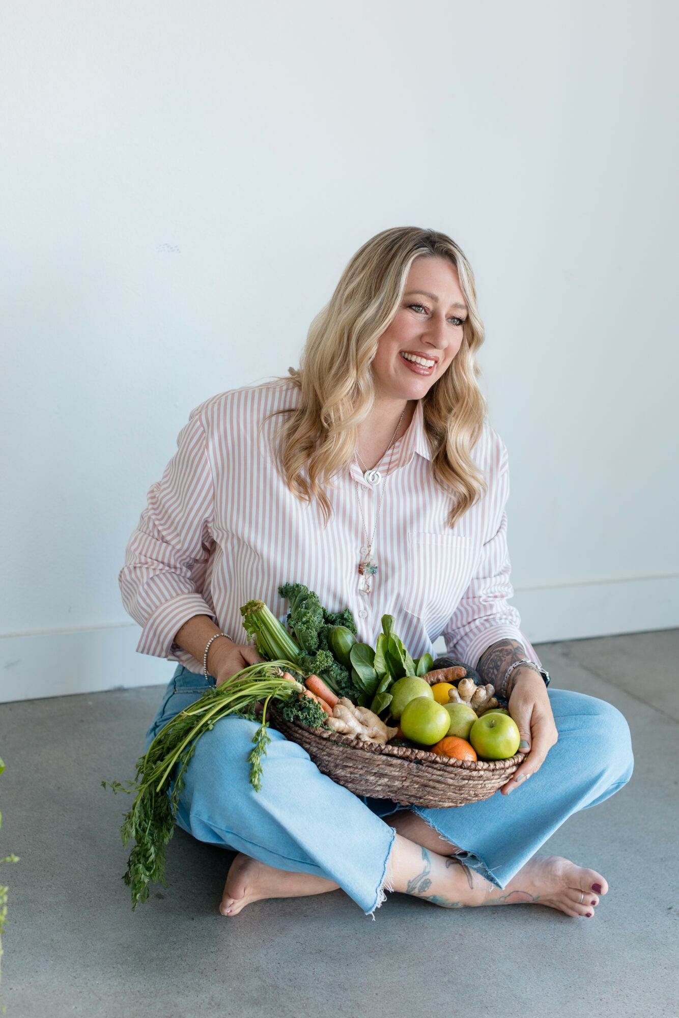 Woman sitting on floor with a basket of vegetables and apples, smiling, against a plain white wall.