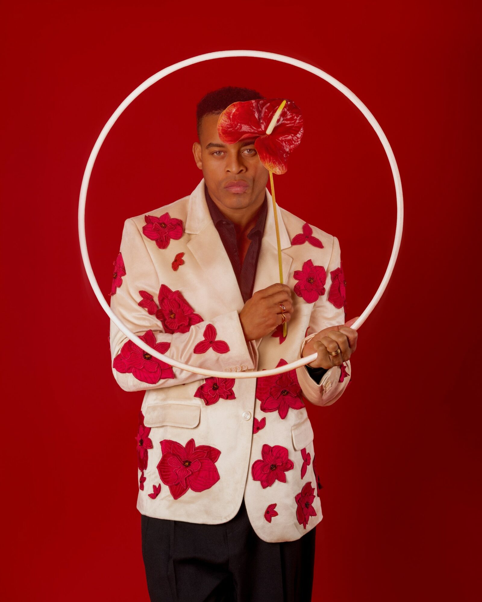 Person holding a hoop with a red feathered accessory, wearing a white jacket with red floral patterns, against a red background.