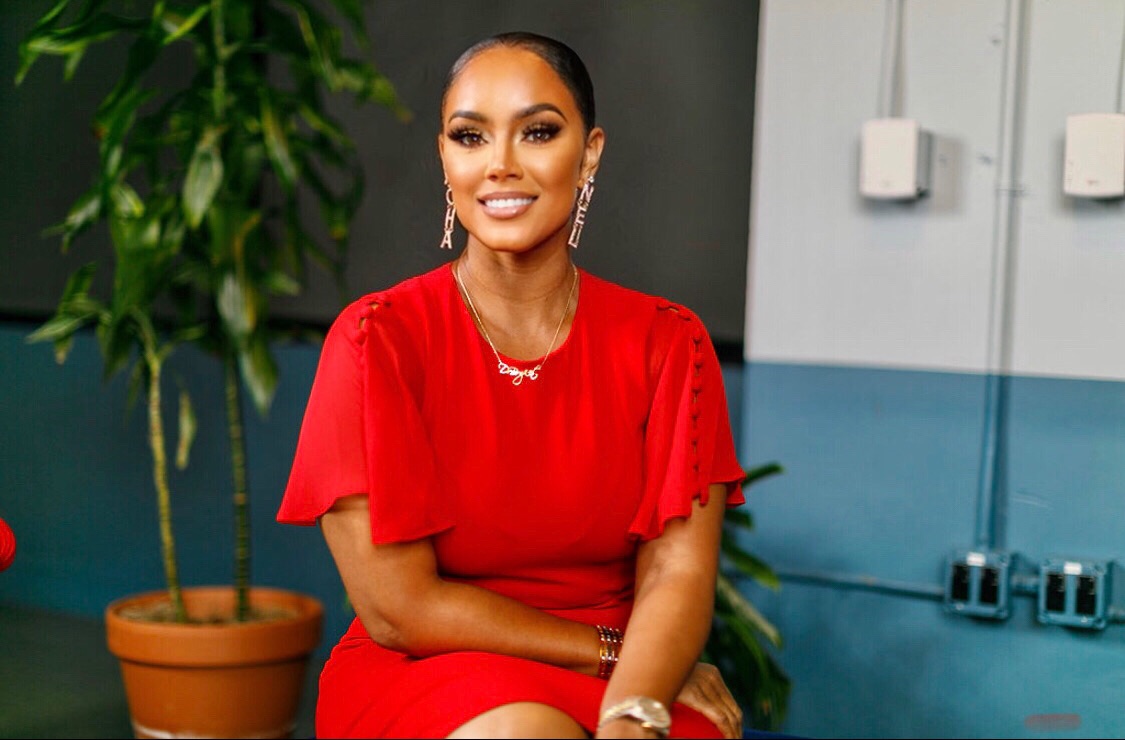 Woman in red dress smiling, sitting indoors with a plant and blue wall in background.