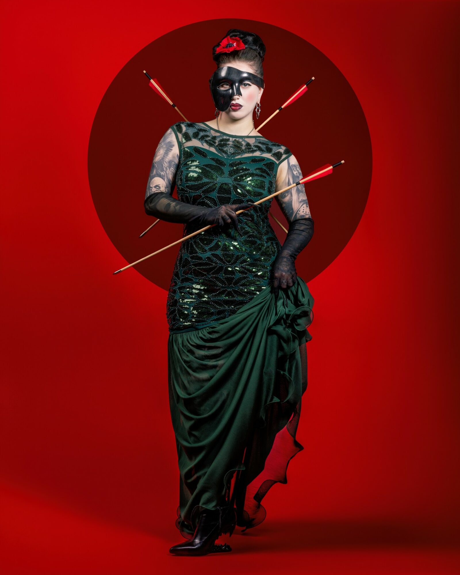Woman in a dark green gown with tattoos, wearing a mask and holding a bow, standing against a red background with arrows.