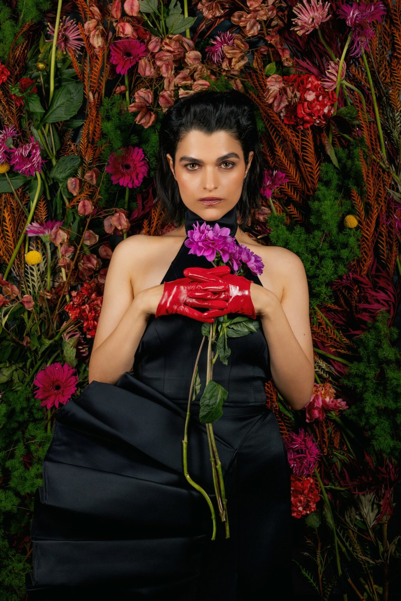 Woman with dark hair and intense expression holding a purple flower, surrounded by colorful flowers and foliage.