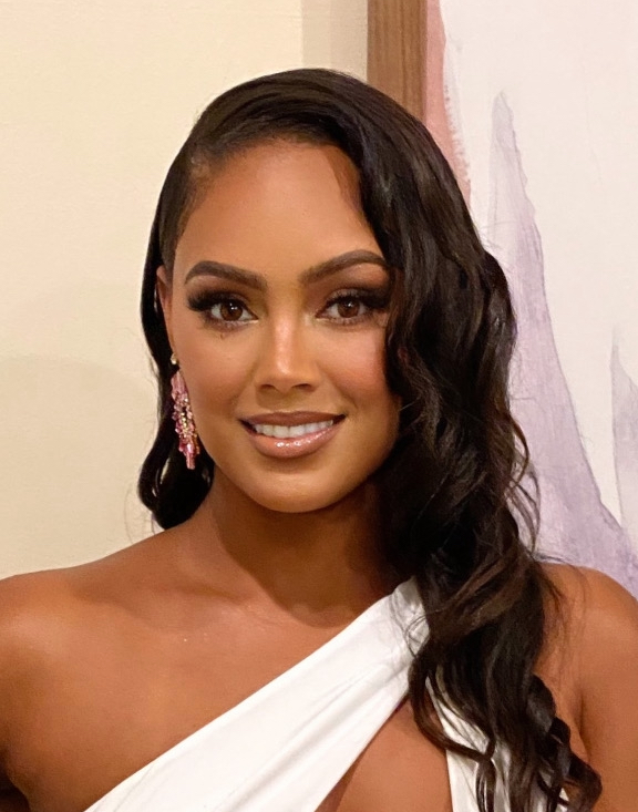 Portrait of a woman with long wavy hair, wearing earrings and a white off-shoulder top, smiling.